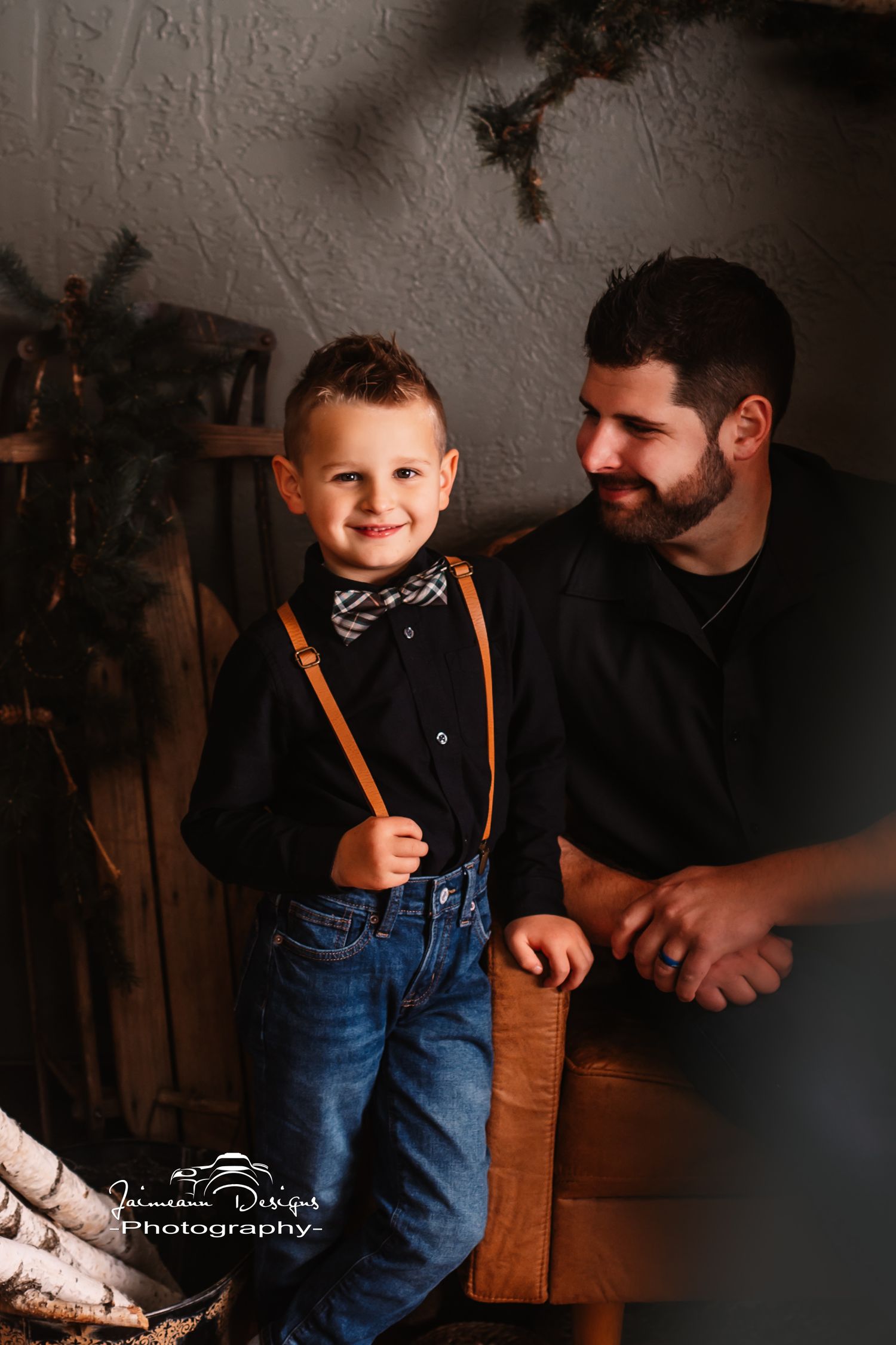 Young boy in suspenders and bowtie poses with adult on leather furniture in dim lighting.