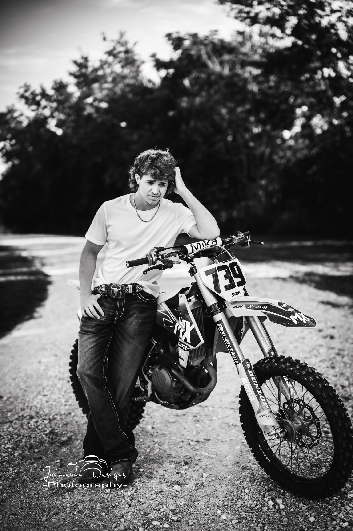 Black and white portrait with dirt bike rider leaning against motorcycle on dirt path.