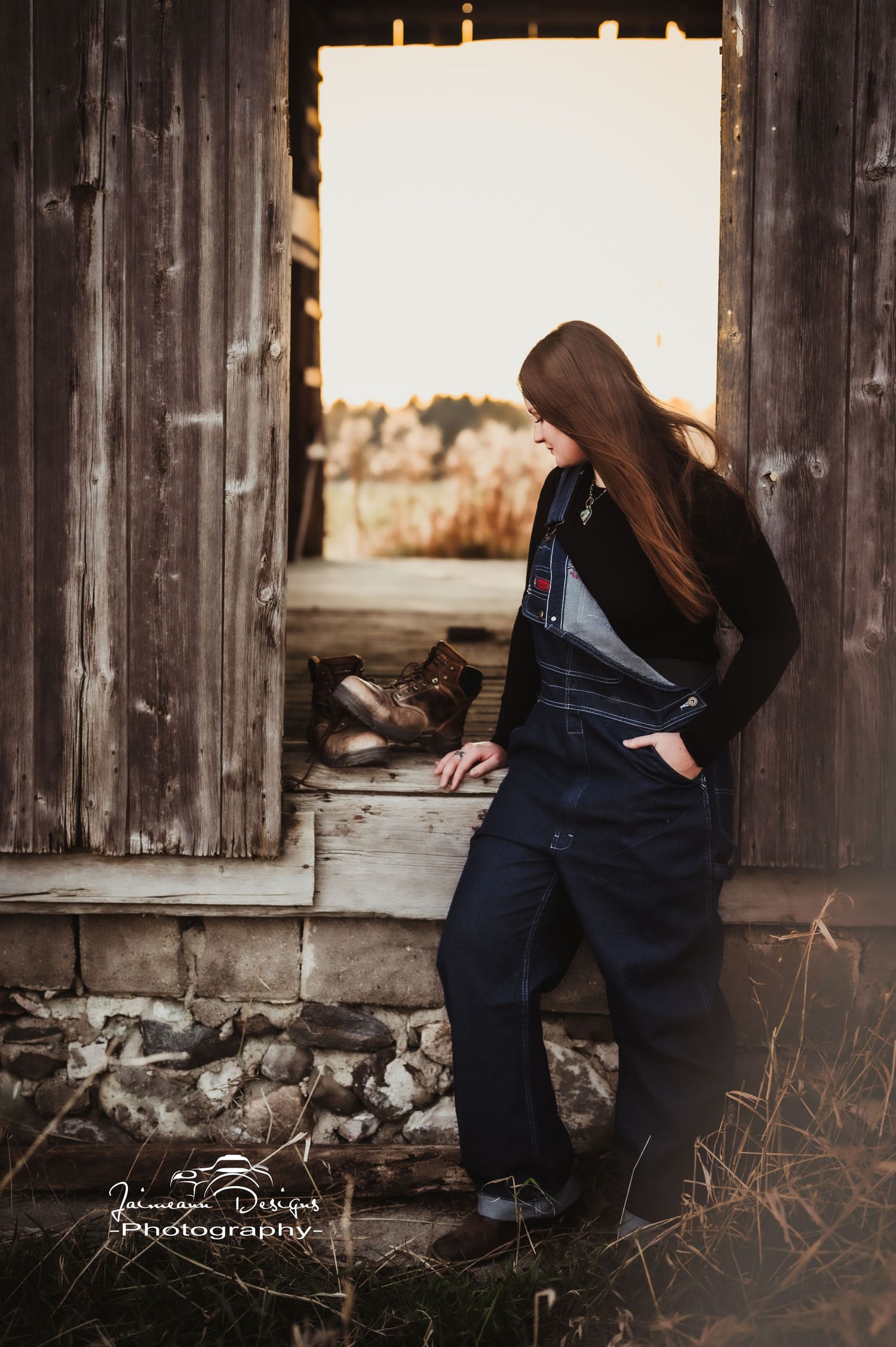 Casual portrait against rustic barn door wearing dark overalls in warm sepia tones.