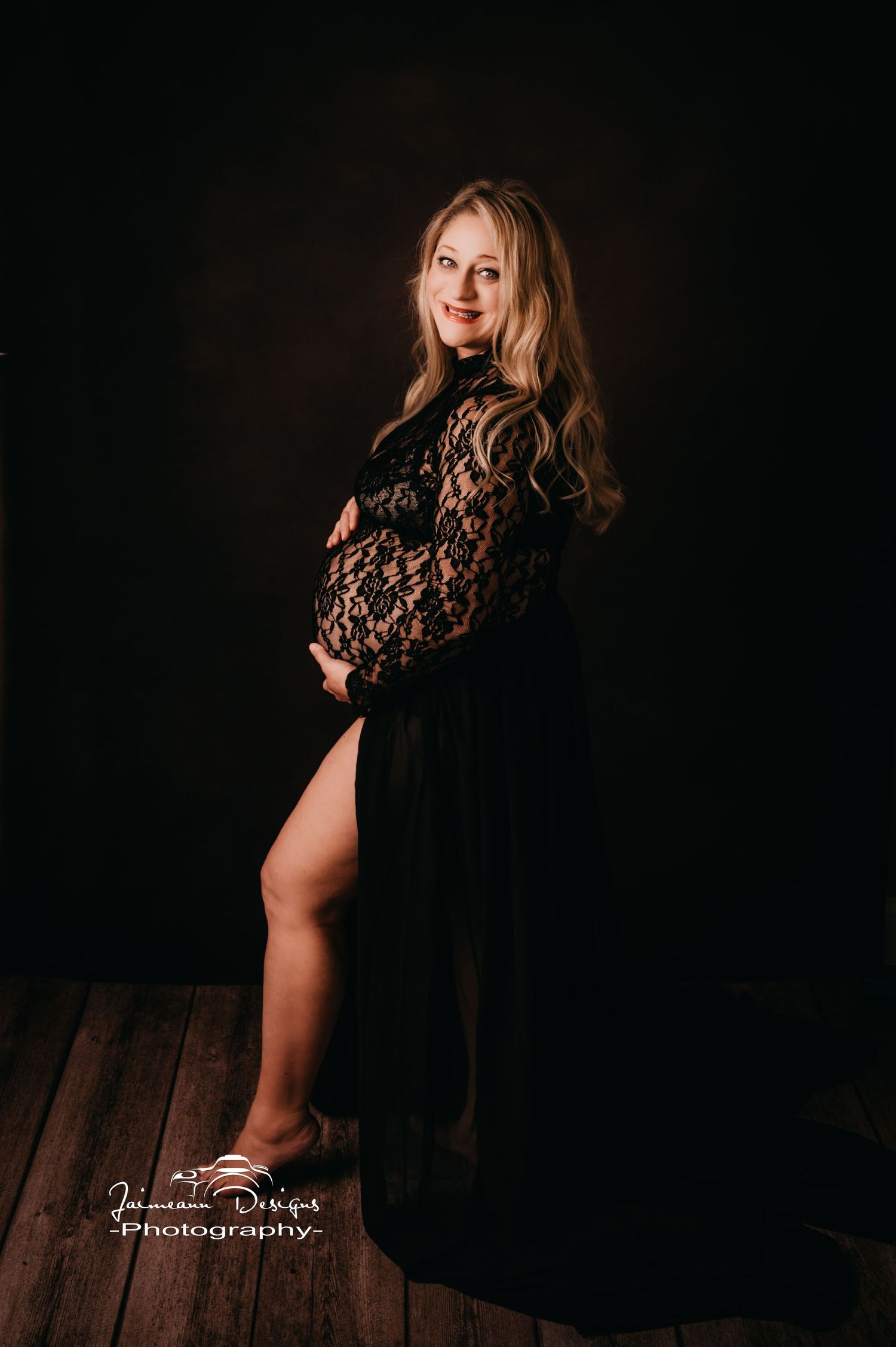 Maternity portrait in black lace dress against dark background with dramatic lighting.