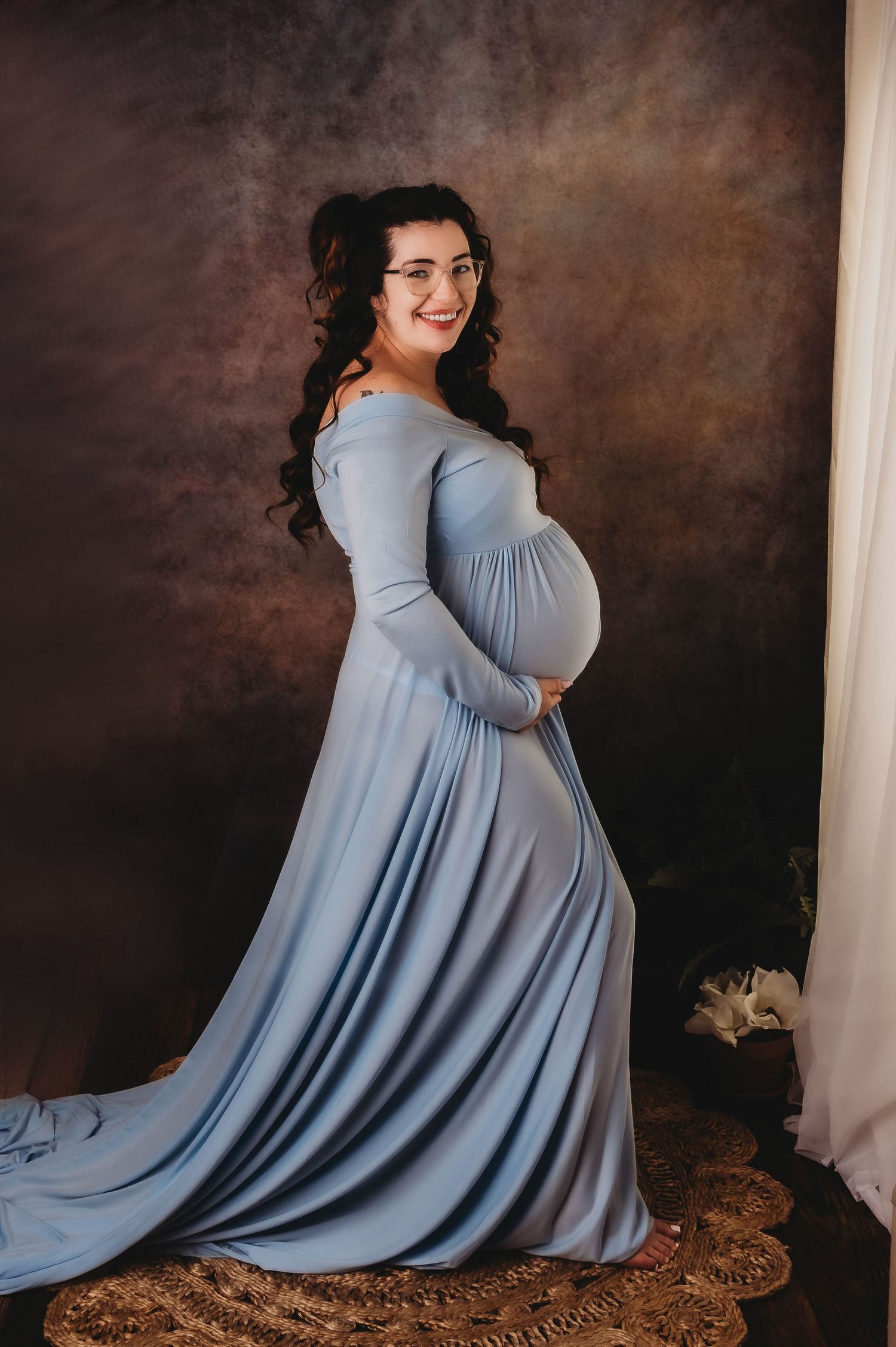 A maternity photo of an expectant mother in a flowing powder blue dress against a dark brown backdrop.