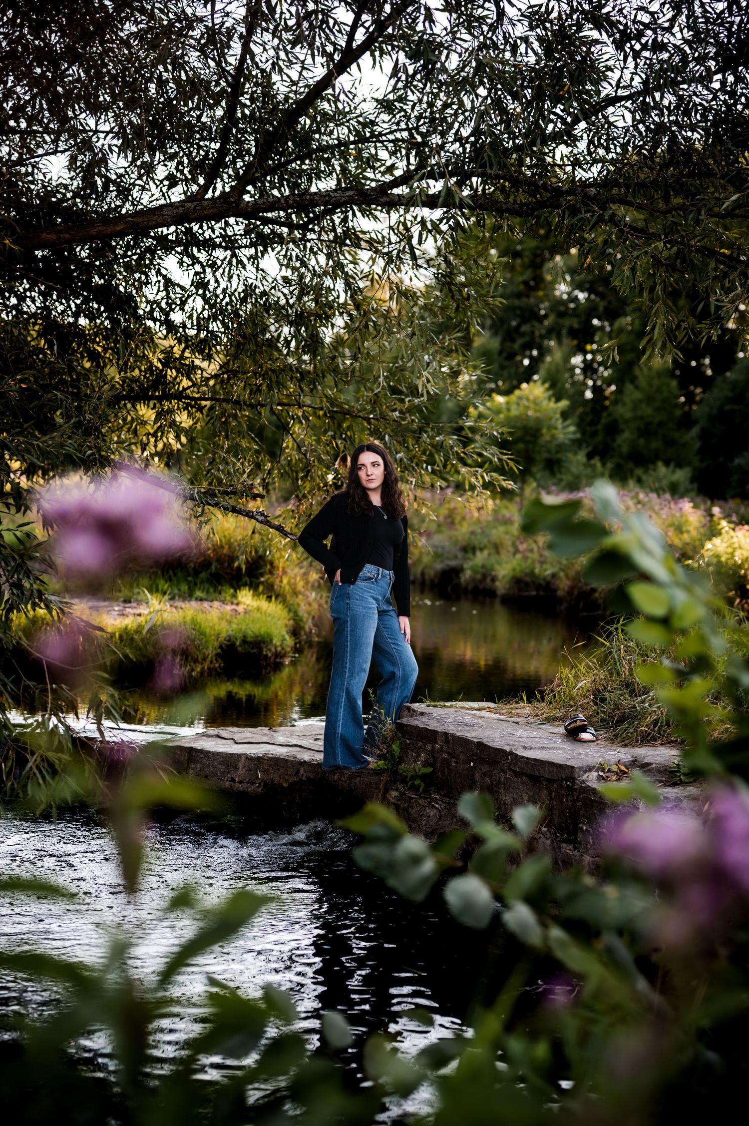 A person stands on a wooden walkway over a tranquil pond, framed by purple flowers and overhanging tree branches at sunset, Kewaskum, WI