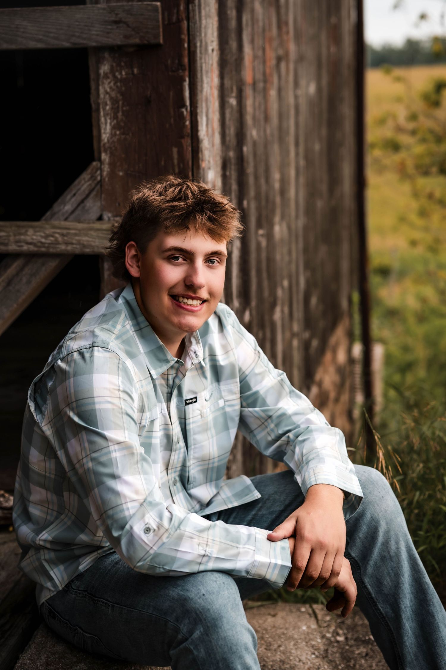 Young man in plaid shirt and jeans smiling while sitting against a rustic wooden barn outdoors.