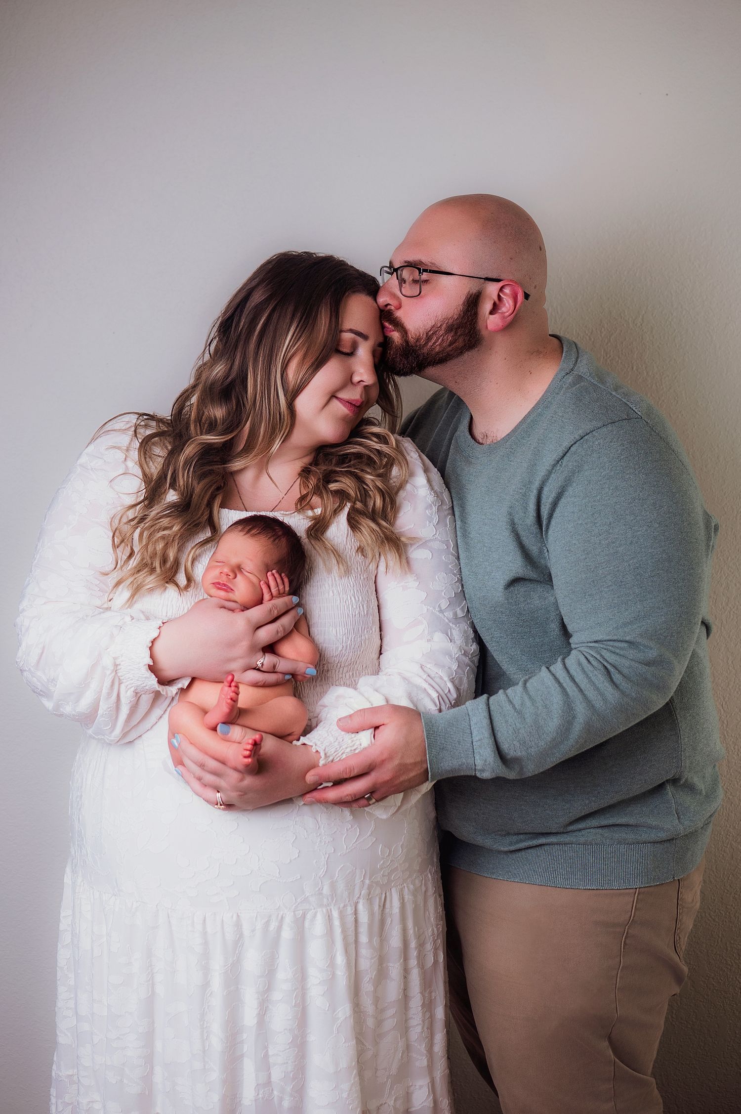 A couple shares a tender moment, the man kissing his partner's temple as she cradles their newborn baby in a white lace dress.
