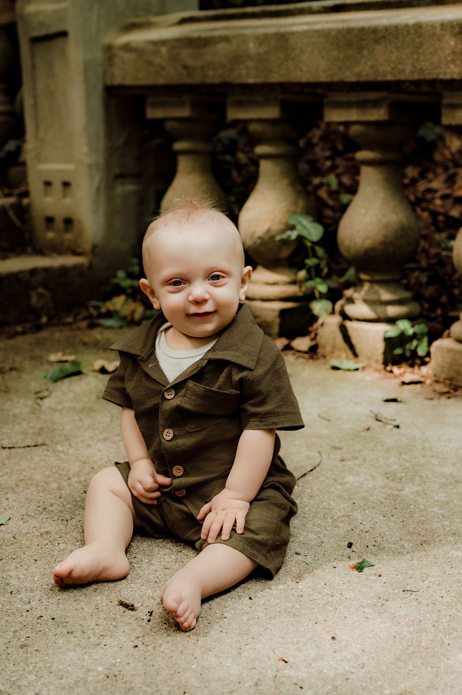 Smiling baby in olive green romper sitting on stone patio near decorative balusters outdoors.