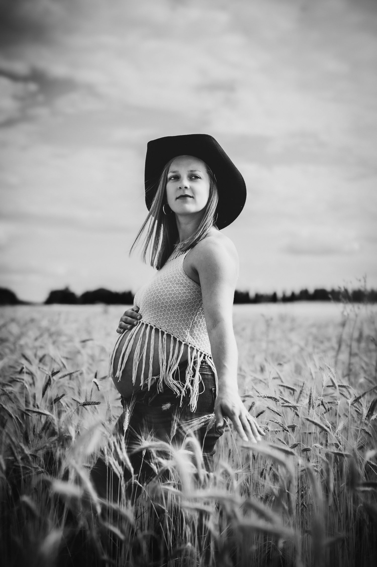 Black and white photo of a pregnant woman in a wide-brim hat standing in a wheat field under a cloudy sky.