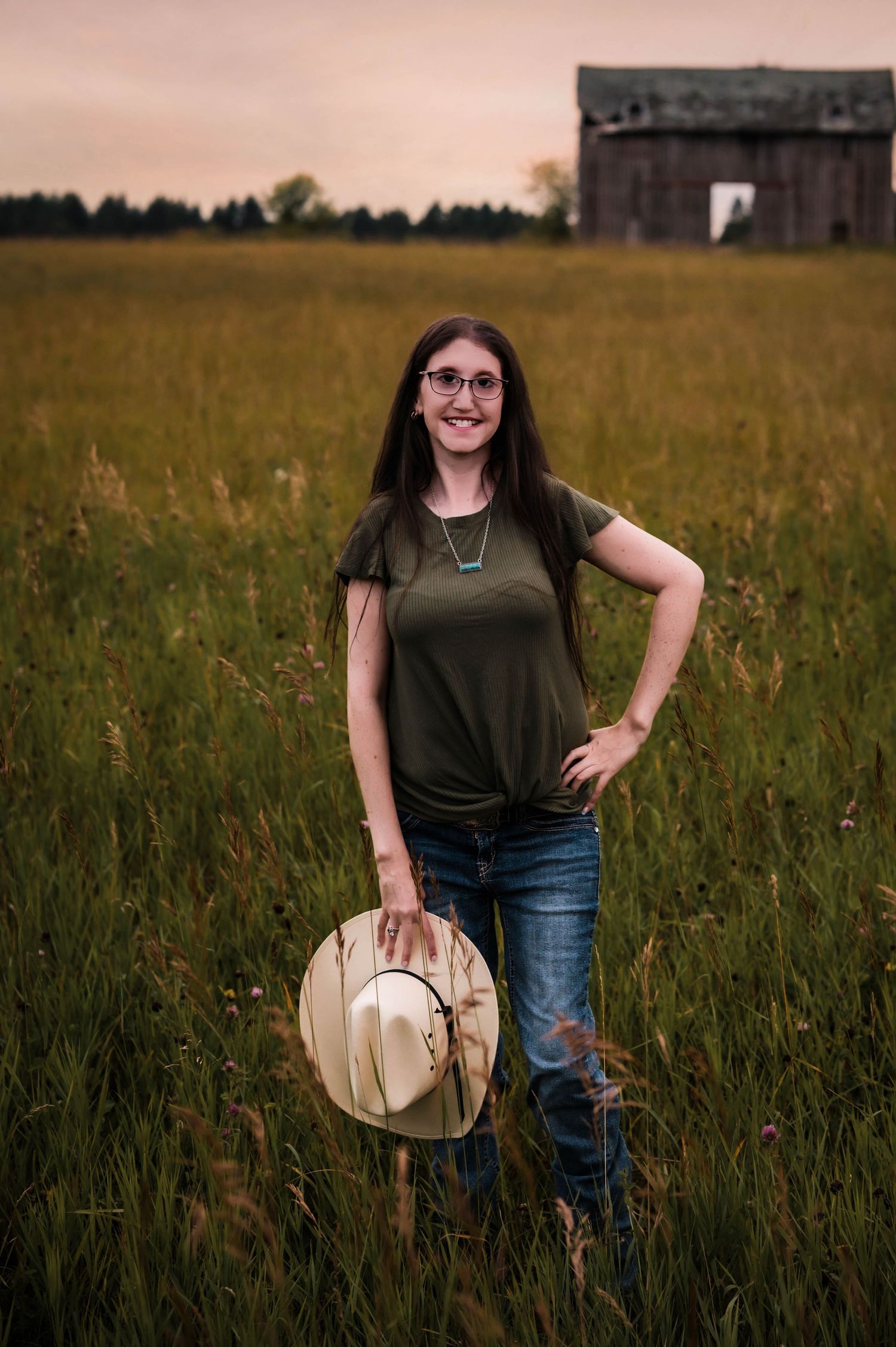 Young woman in green shirt and jeans holds cowboy hat in a field with rustic barn at sunset.