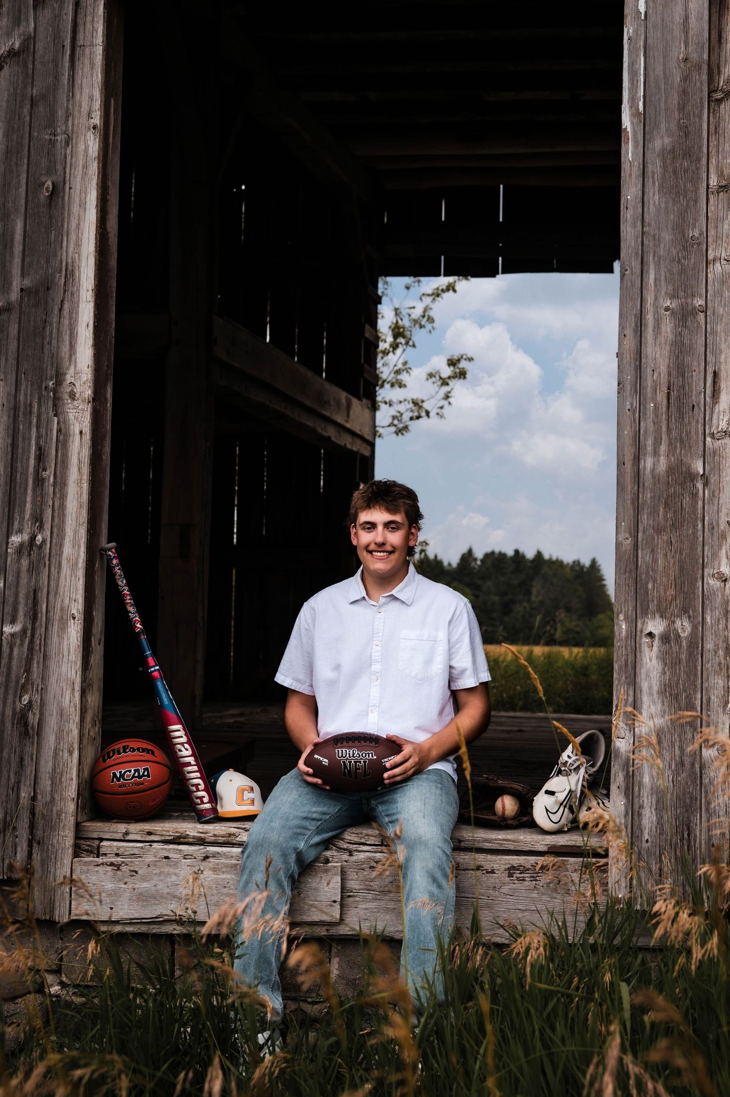 A smiling teen holds a football in a rustic barn doorway, surrounded by sports equipment including a basketball and baseball bat.