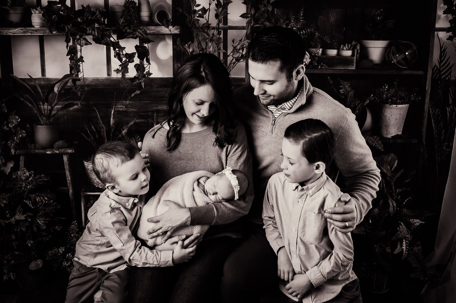 Black and white family portrait showing parents with children and newborn in a cozy indoor setting.