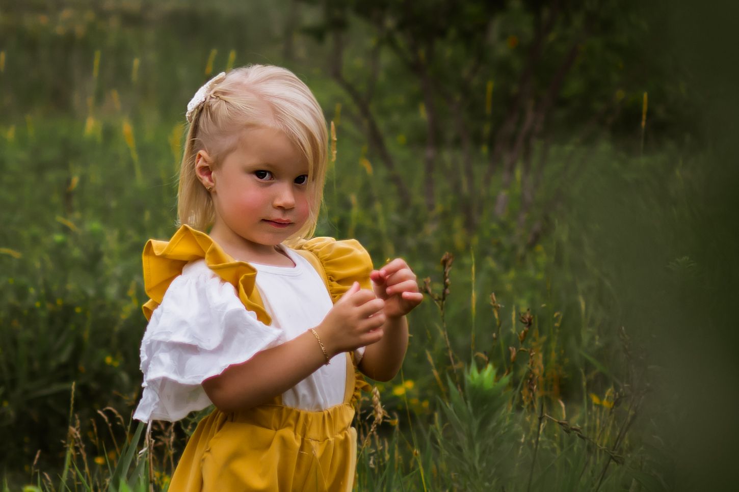 Child in yellow overalls and white blouse stands in wildflower meadow during golden hour.