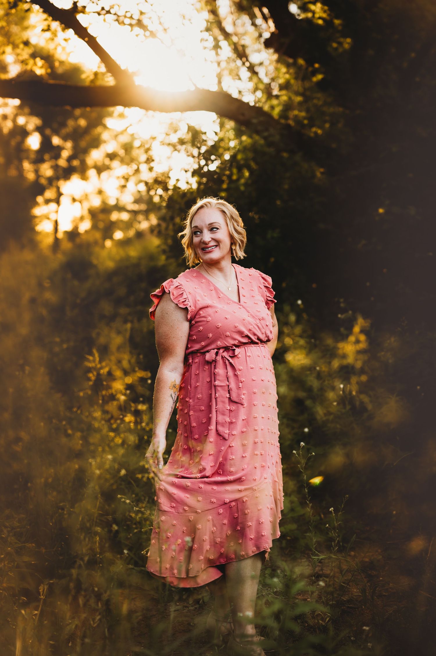 A person in a pink dress smiles in a sunlit forest setting during golden hour.