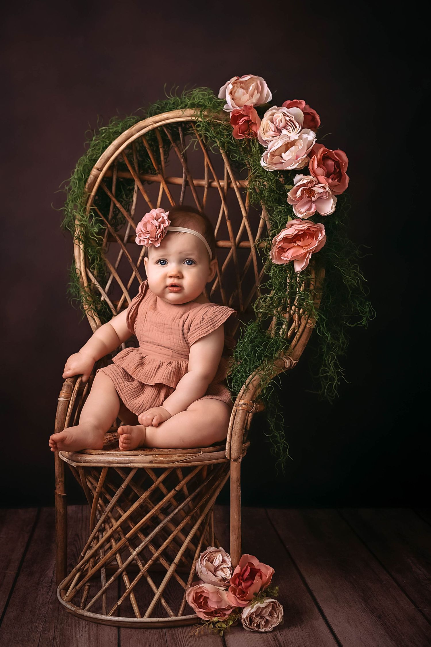 A wicker chair decorated with pink flowers and greenery serves as a beautiful backdrop for a baby portrait session.