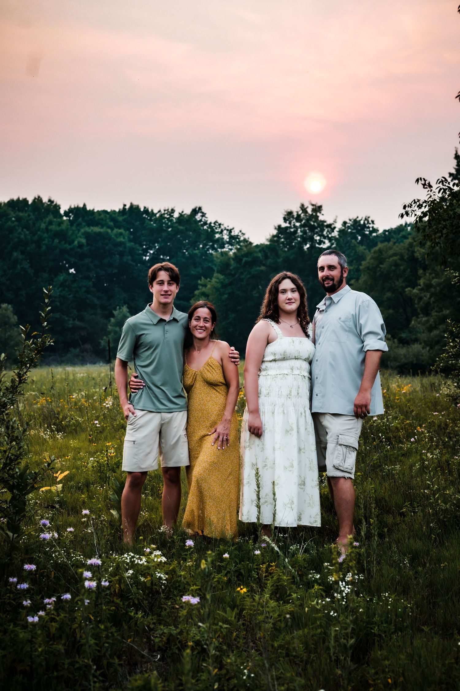 Family of four posing in a wildflower meadow at sunset with lush green trees in the background.