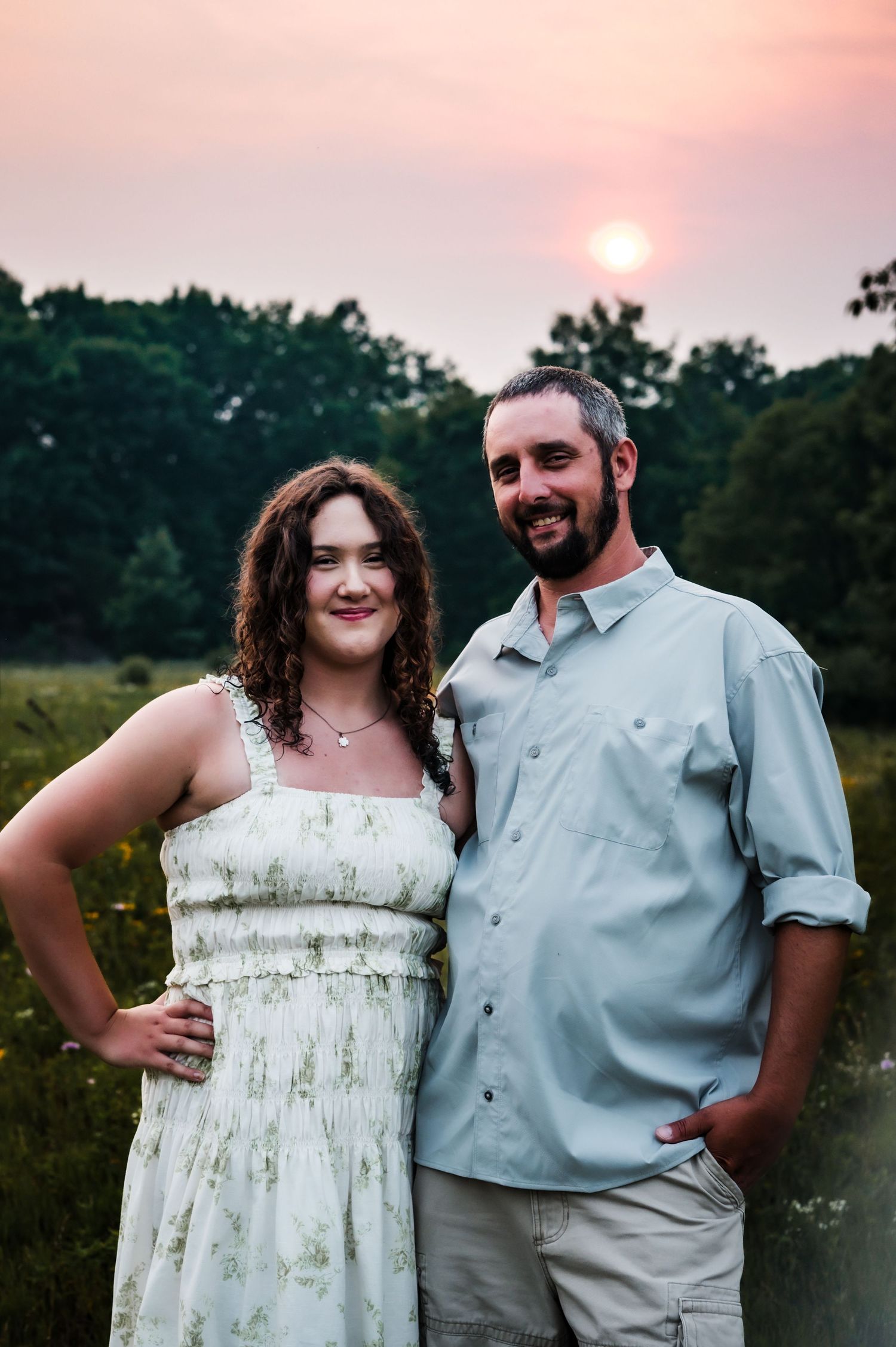 A smiling couple poses together at sunset in a field, woman in floral dress and man in light blue shirt.
