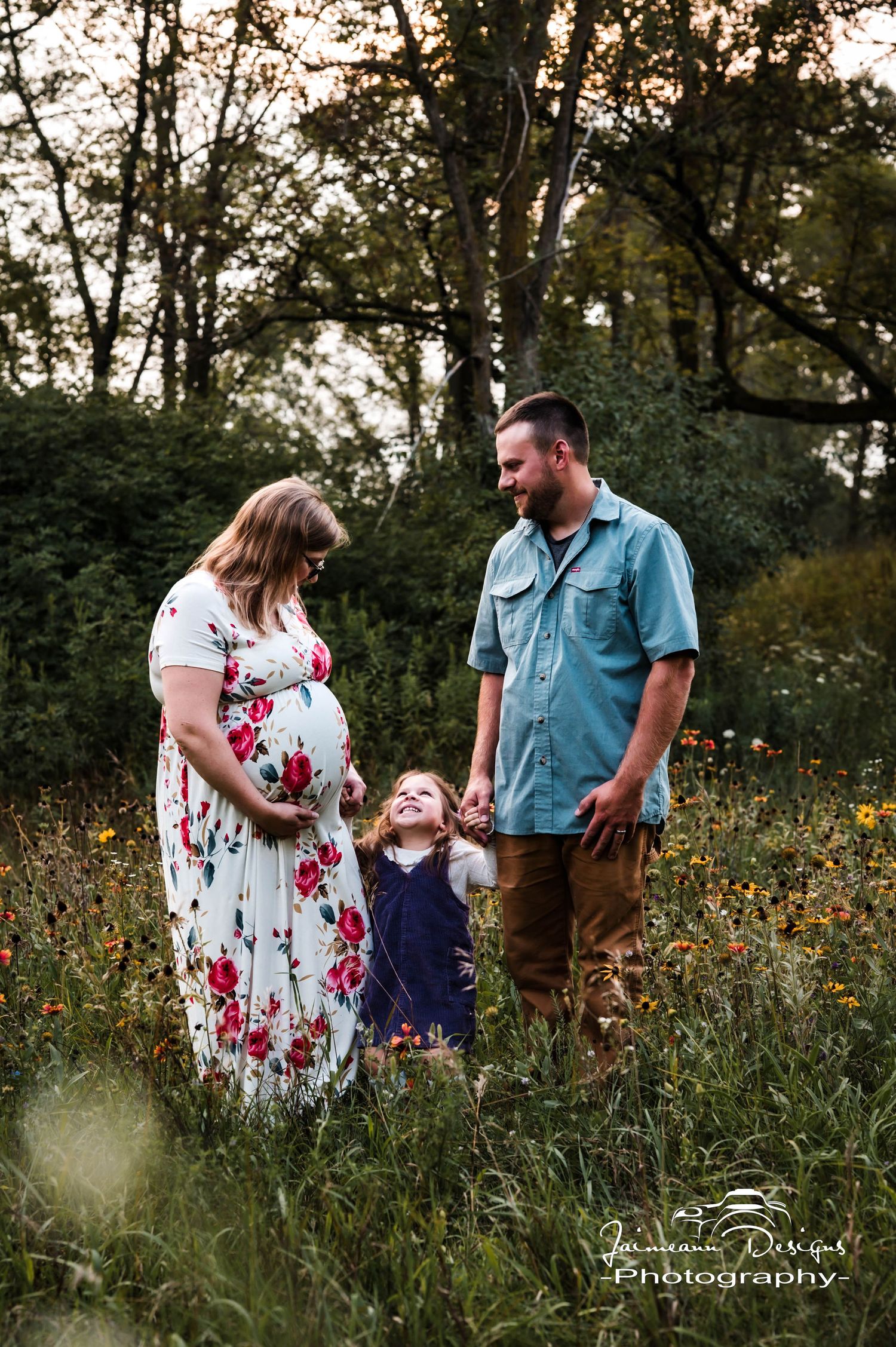 Pregnant woman in floral dress with partner and child in wildflower field during golden hour maternity photography session.