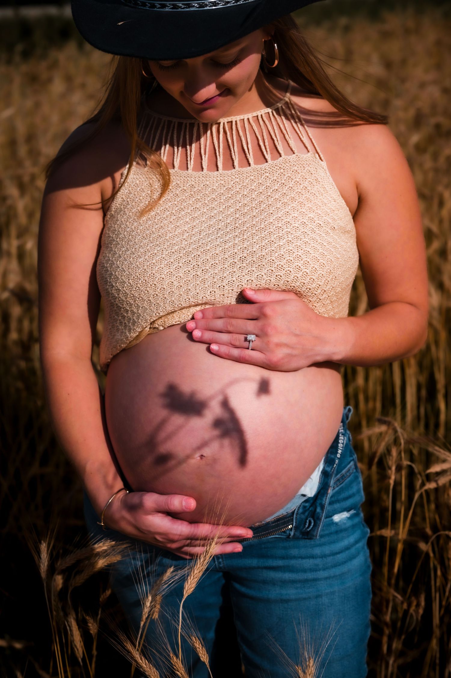 Pregnant woman in cowboy hat and crop top cradling baby bump in golden wheat field during sunset.