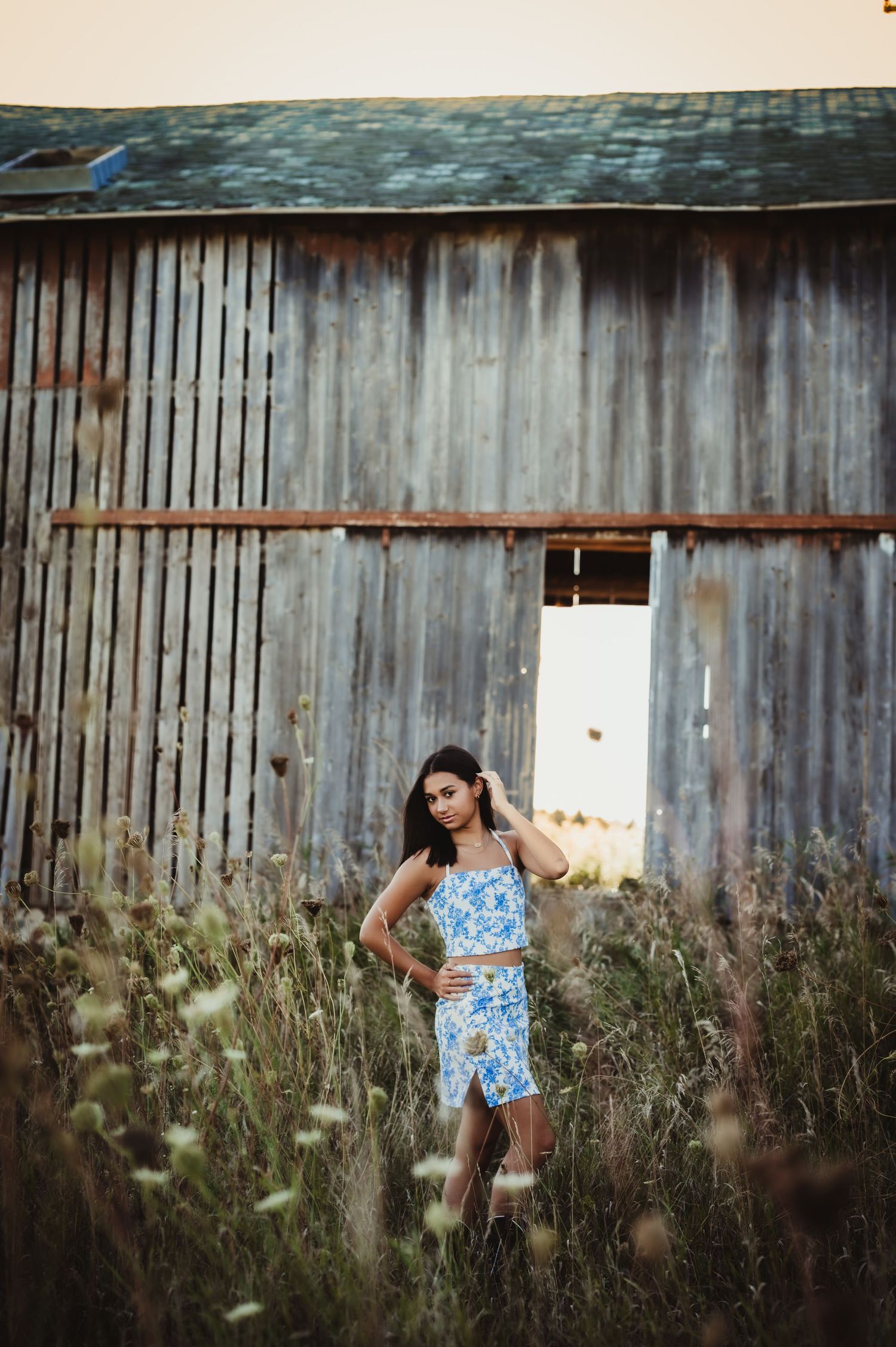 Person in blue floral dress poses in front of rustic barn with wildflowers.