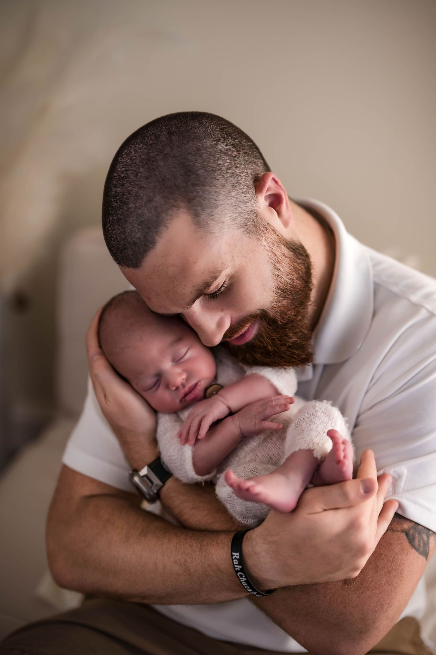 Bearded father in a white polo tenderly cradles his sleeping newborn baby against his chest in a warm, intimate newborn portrait.