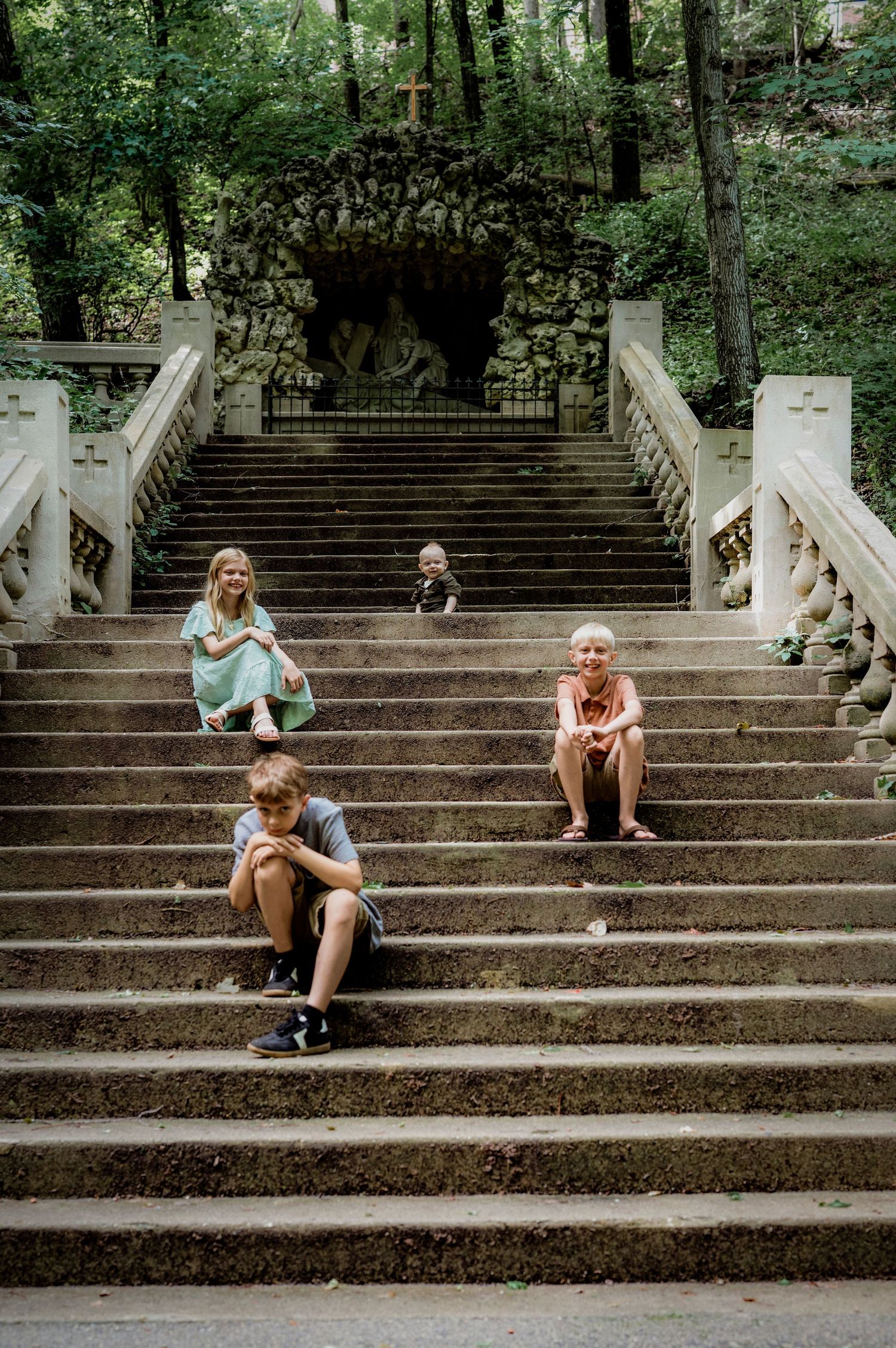 Four children sitting on ornate stone stairs surrounded by lush green trees near a decorative grotto.