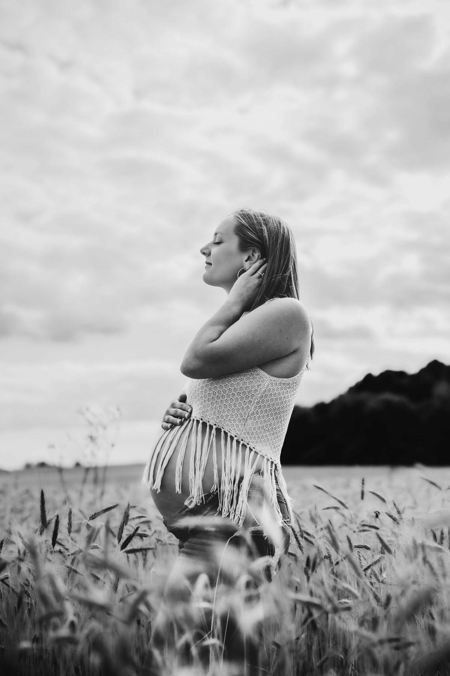 Black and white photo of a pregnant woman standing in a wheat field, cradling her belly with eyes closed.