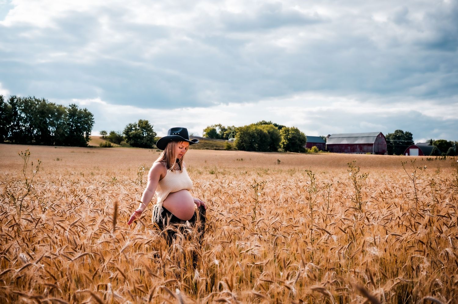 Pregnant woman in a wheat field wearing a black hat and white top, with a red barn and trees in the background.
