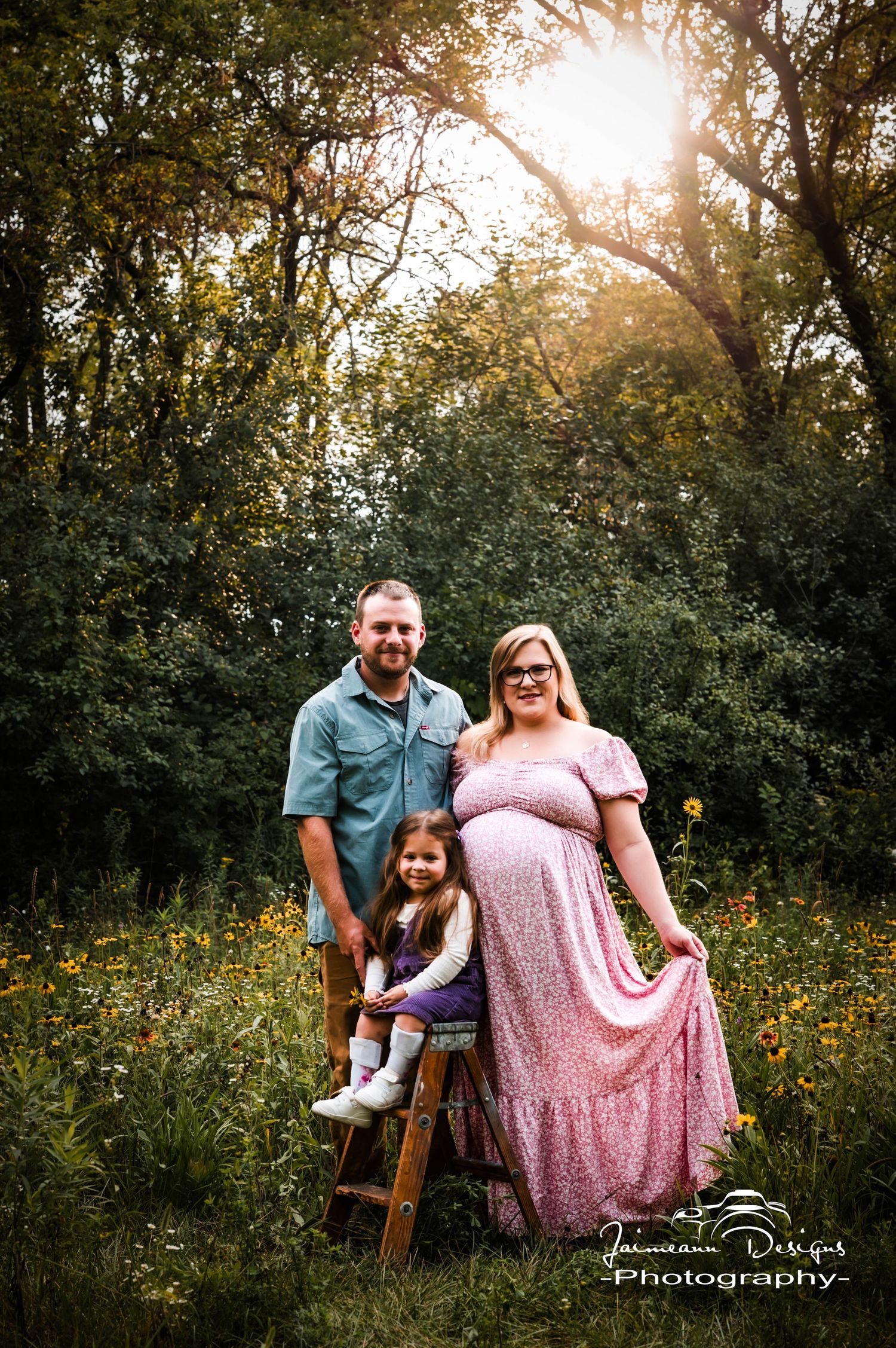 Pregnant woman in pink dress poses with husband and young daughter in a sunlit wildflower meadow for maternity photos.