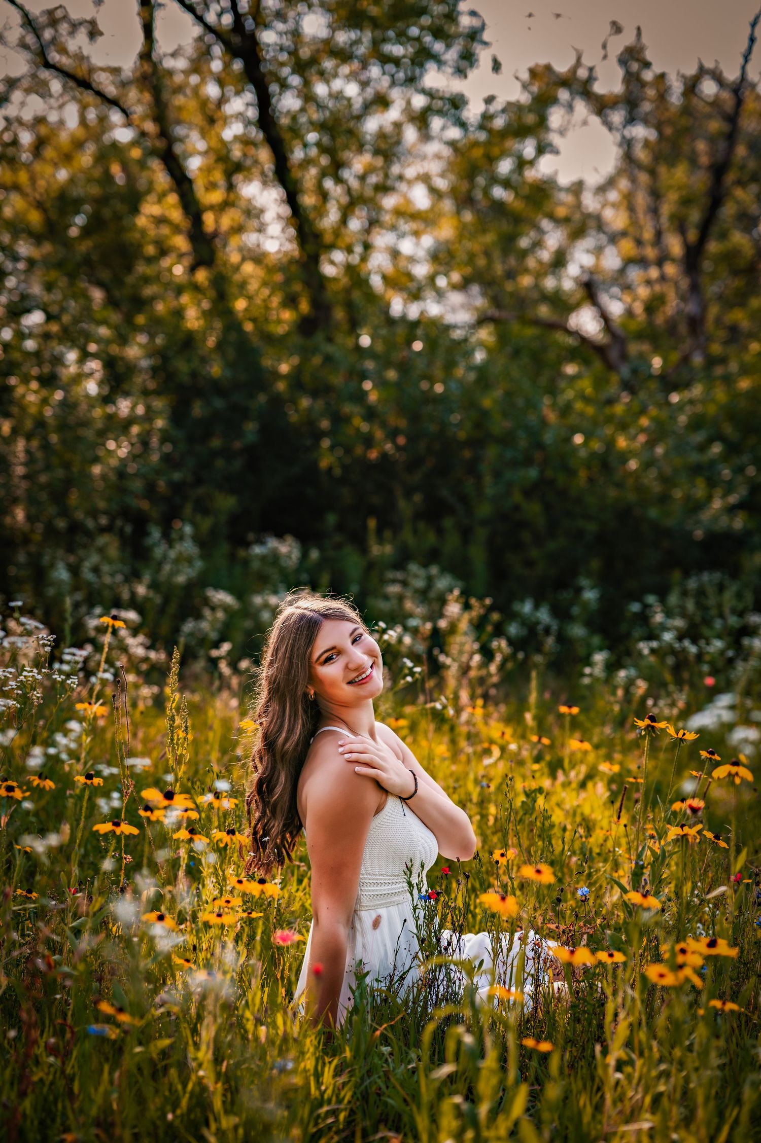 A person in a white dress stands among wildflowers in a golden meadow at sunset with trees in the background.