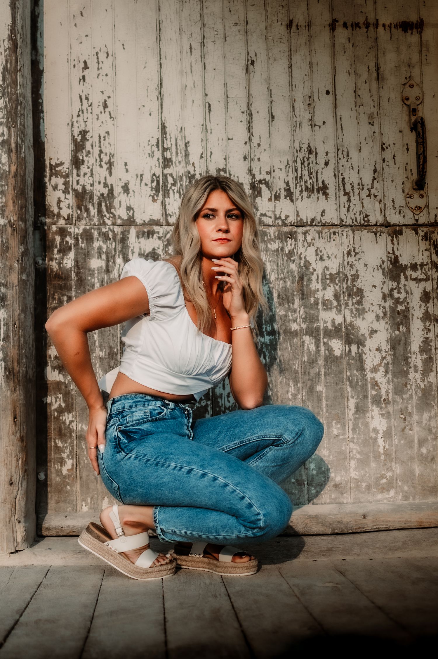 Person in white top and jeans crouches against weathered wall in artistic pose.
