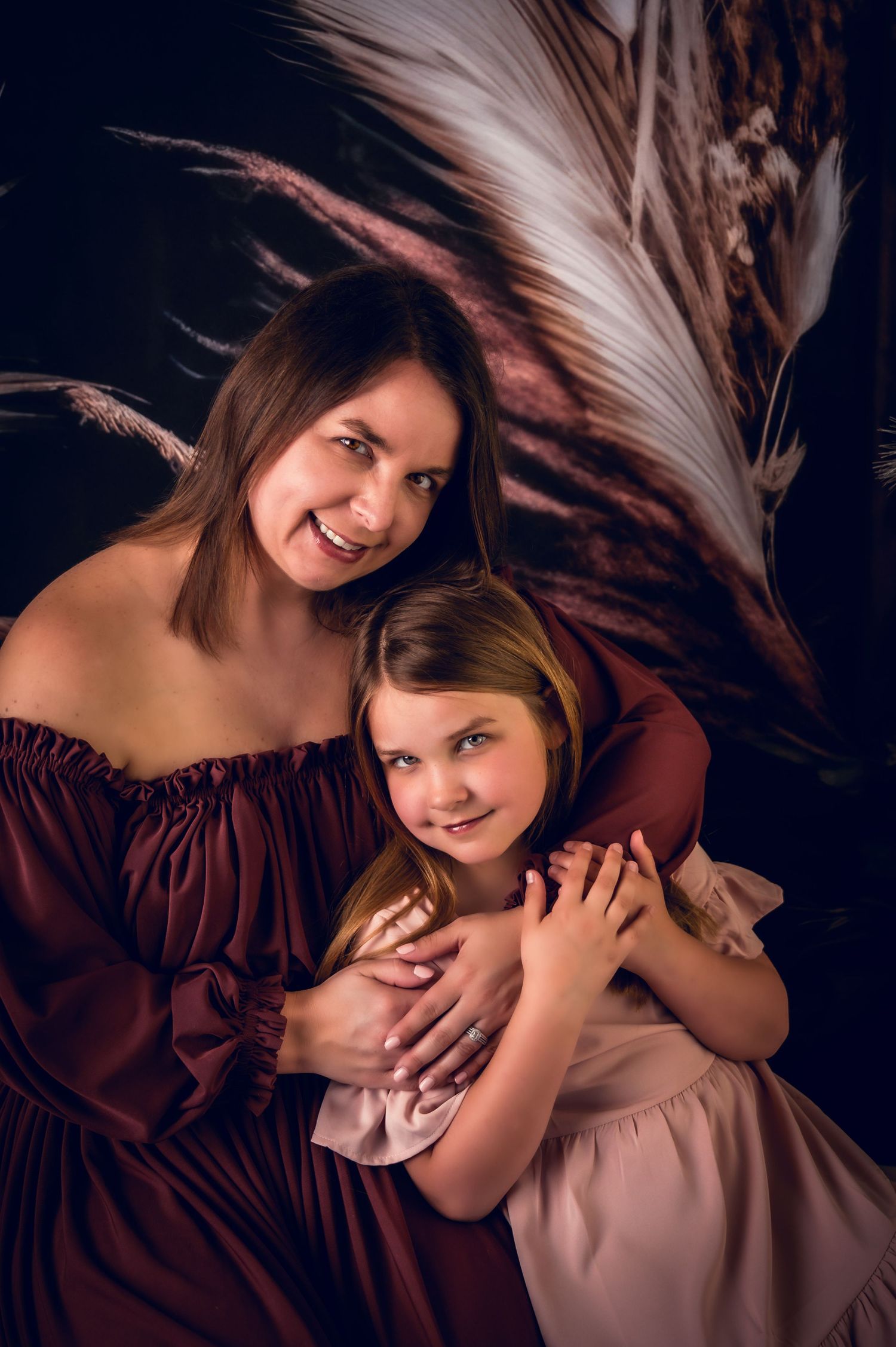 Loving embrace captured in burgundy and pink dresses against feathered background.