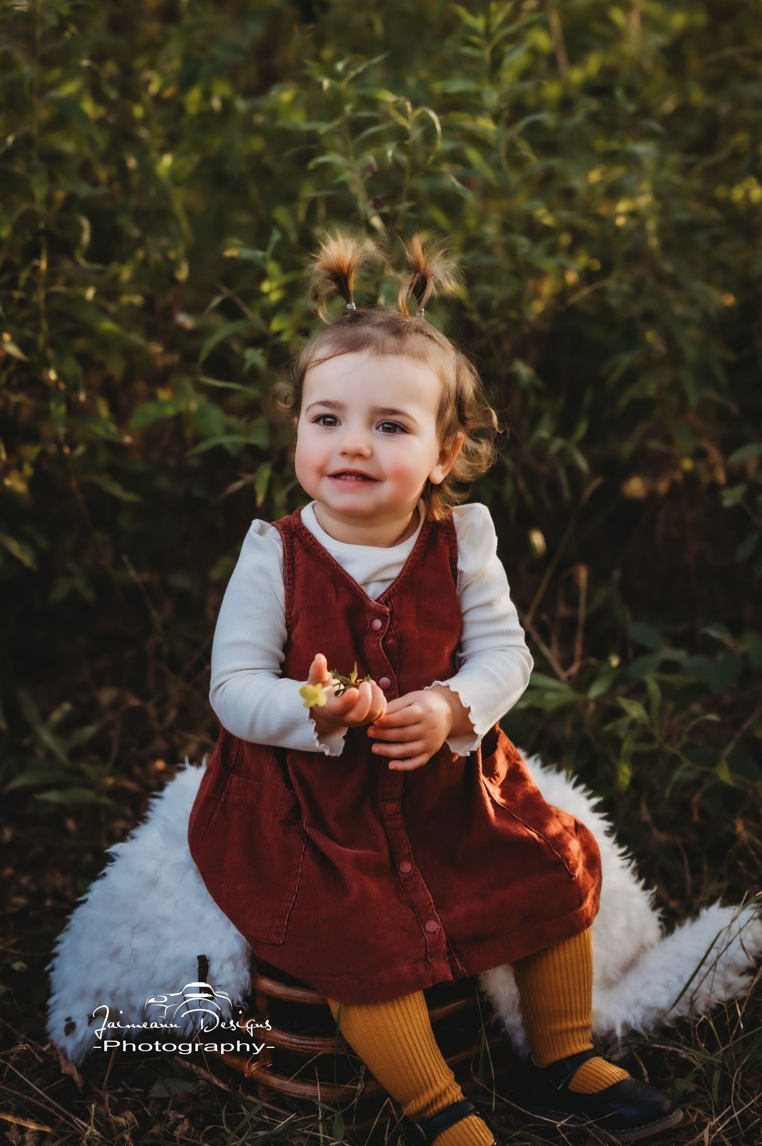 A toddler in a burgundy dress and yellow tights sits on a white surface outdoors.