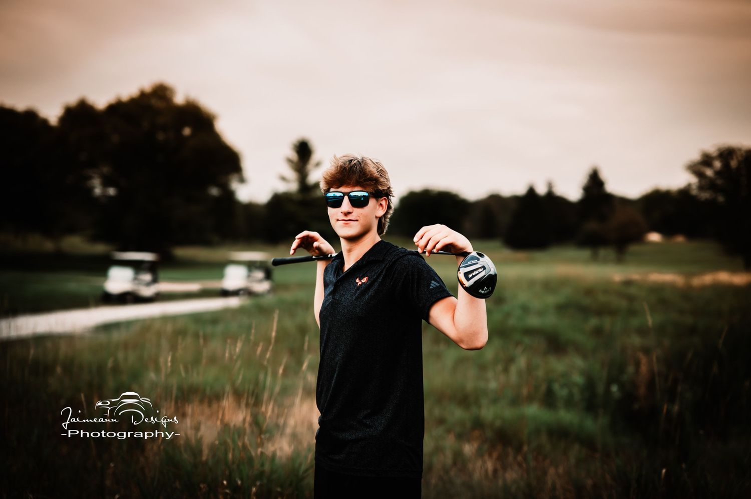 Golfer poses with club over shoulder against sunset at golf course with cart in background.