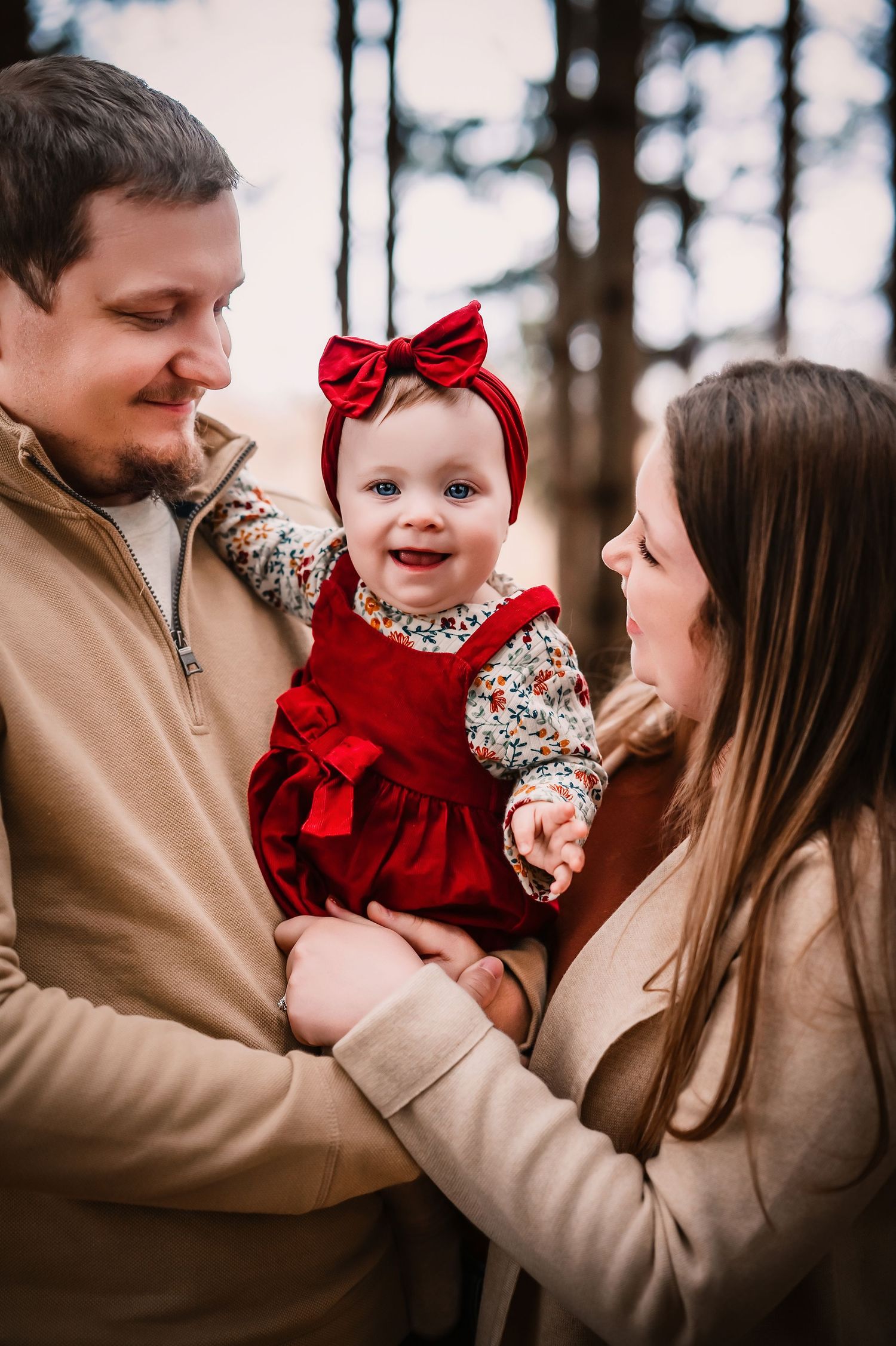 Happy baby in red dress with bow held by smiling parents outdoors in a forest setting.