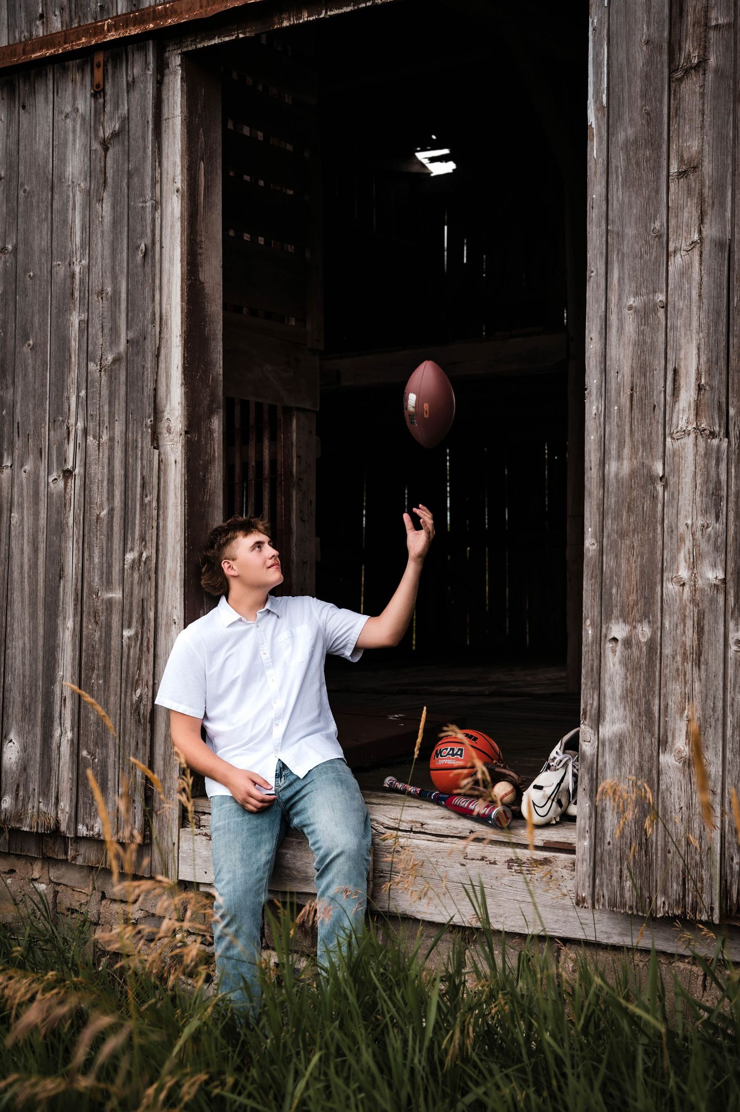 Young man in white shirt and jeans tossing a football outside a rustic wooden barn with sports equipment nearby.