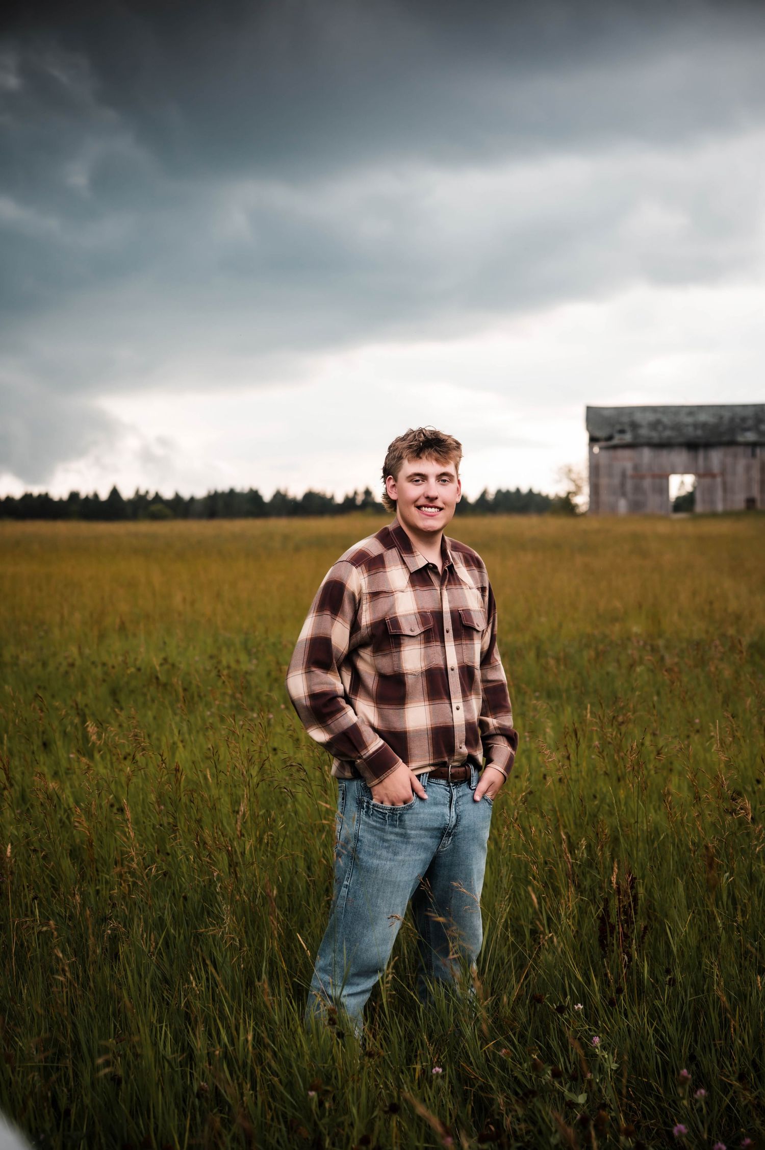 Young man in plaid shirt and jeans smiling in a green field with a rustic barn and stormy sky.