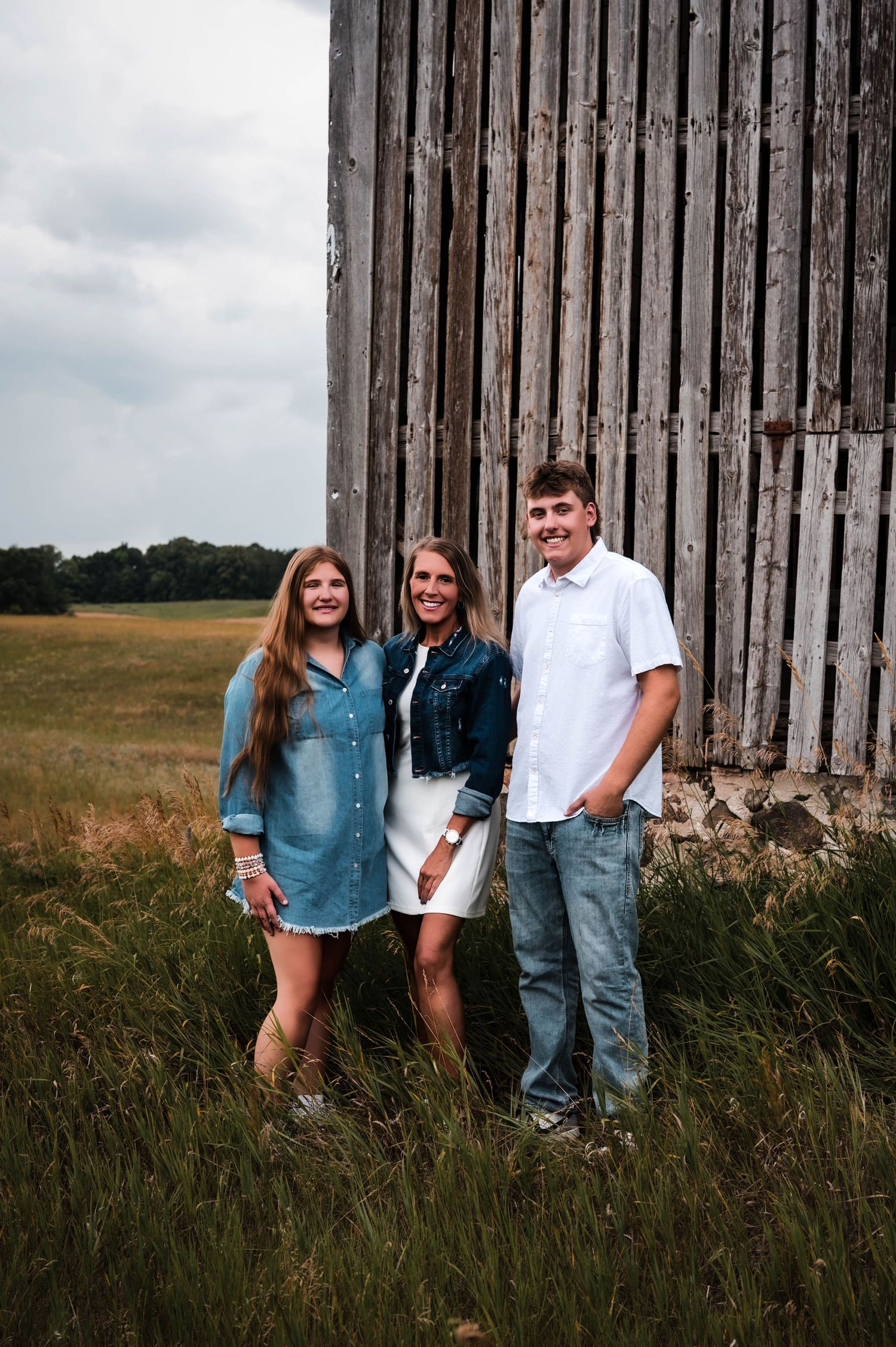 Three smiling people pose beside a rustic wooden barn in a green field, wearing casual denim outfits on a cloudy day.