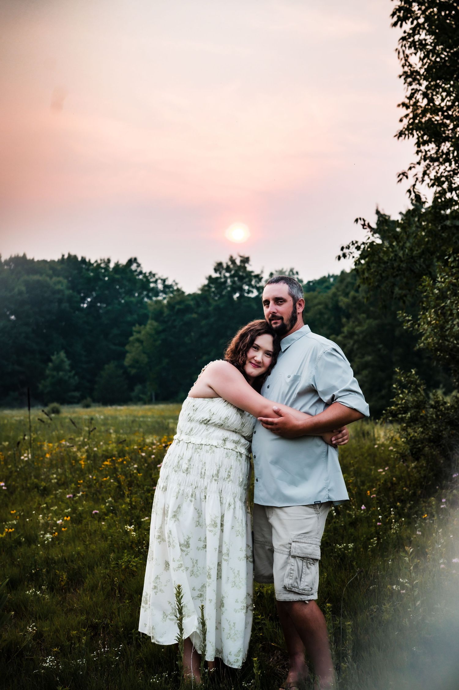 Couple embracing at sunset in a wildflower meadow, woman in white dress hugging man in gray shirt, trees in background.
