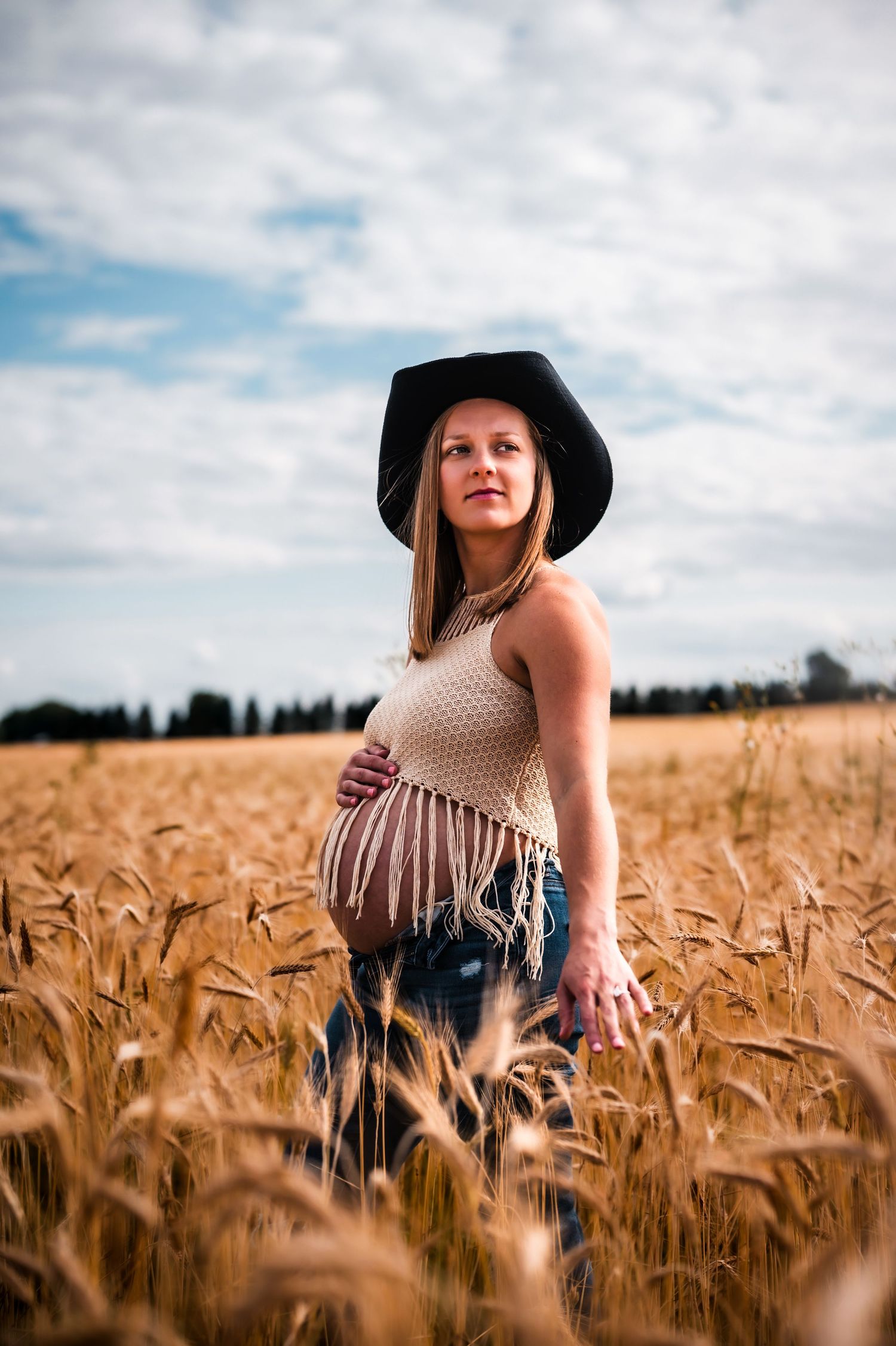 Pregnant woman in black cowboy hat and crochet top stands in golden wheat field under cloudy sky.