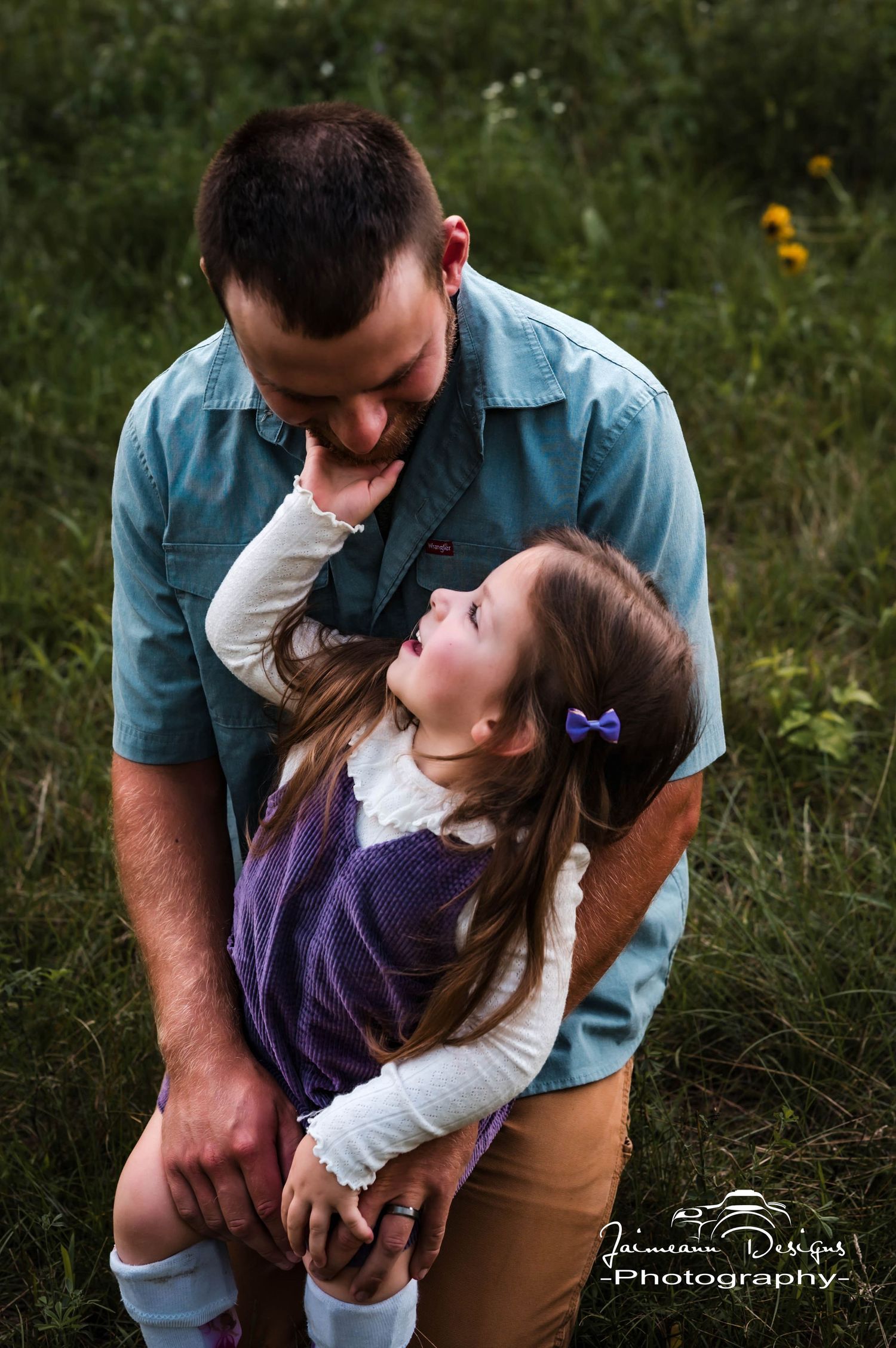 A father kneels in a green field as his young daughter in a purple dress reaches up to touch his face tenderly.