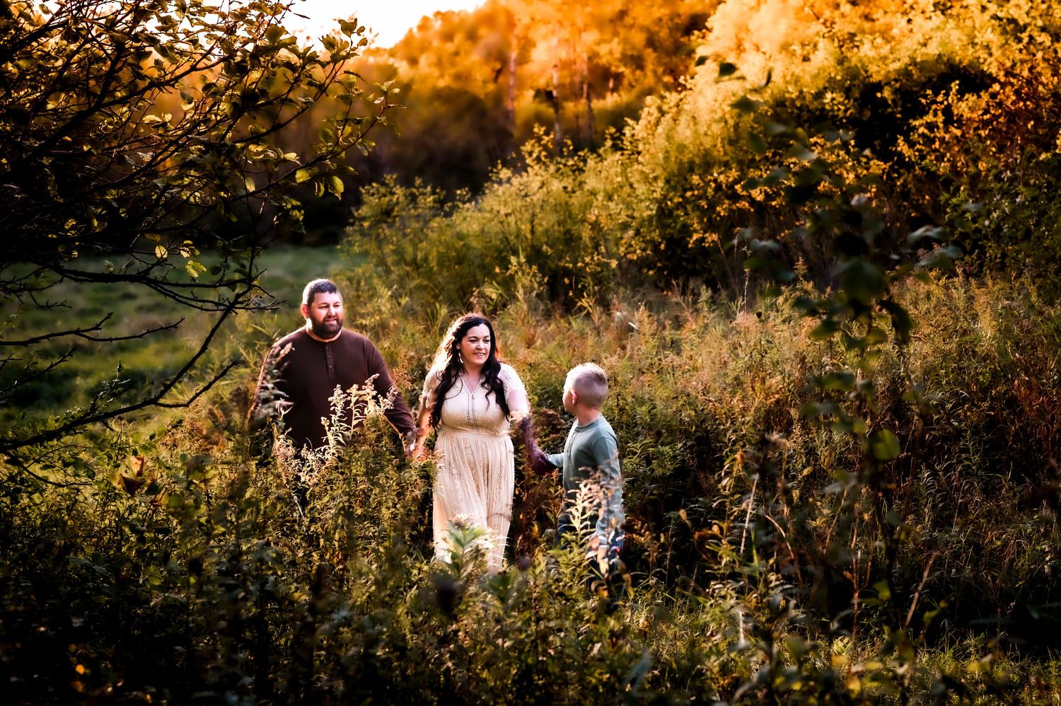 Family walking through golden autumn meadow with lush foliage, warm sunset light illuminating their joyful outdoor portrait.