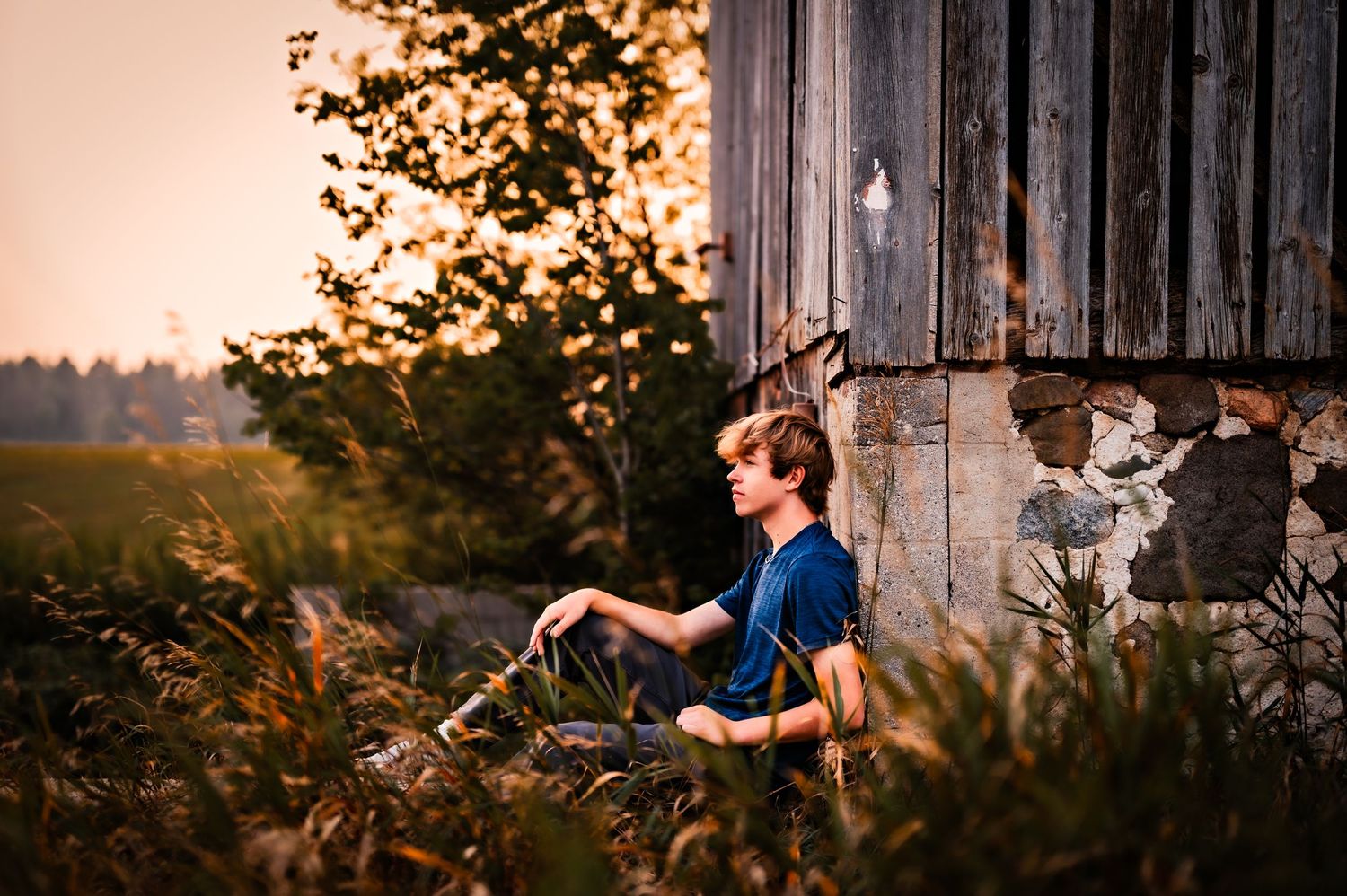 A couple sits together against a rustic barn wall during golden hour sunset in a romantic outdoor setting.