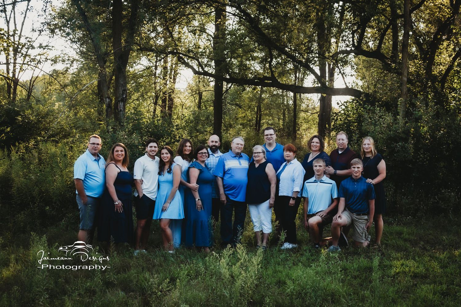 Extended family portrait in coordinating blue outfits standing together in a sunlit forest clearing.