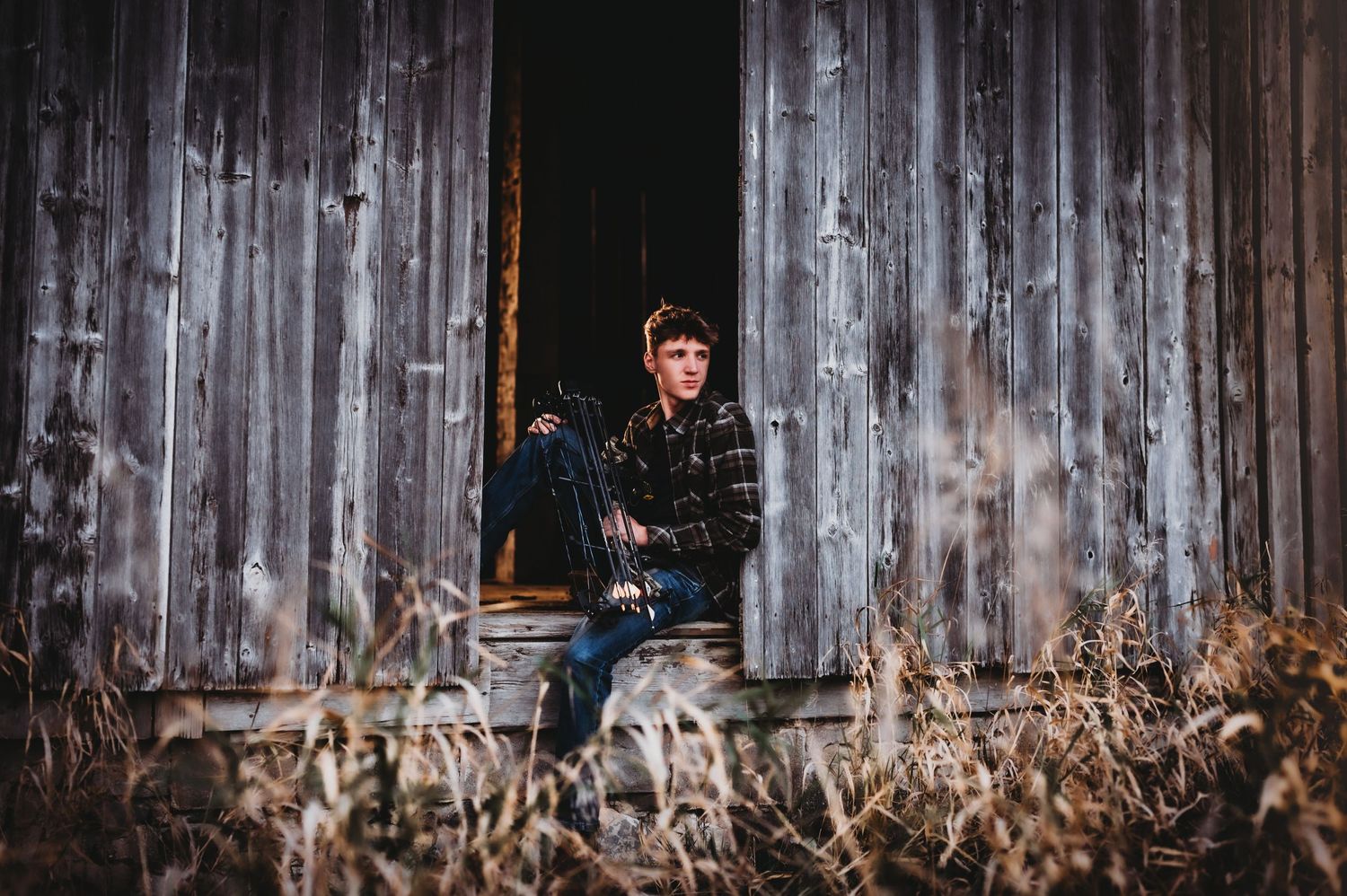 Figure stands at weathered barn entrance surrounded by tall grass.
