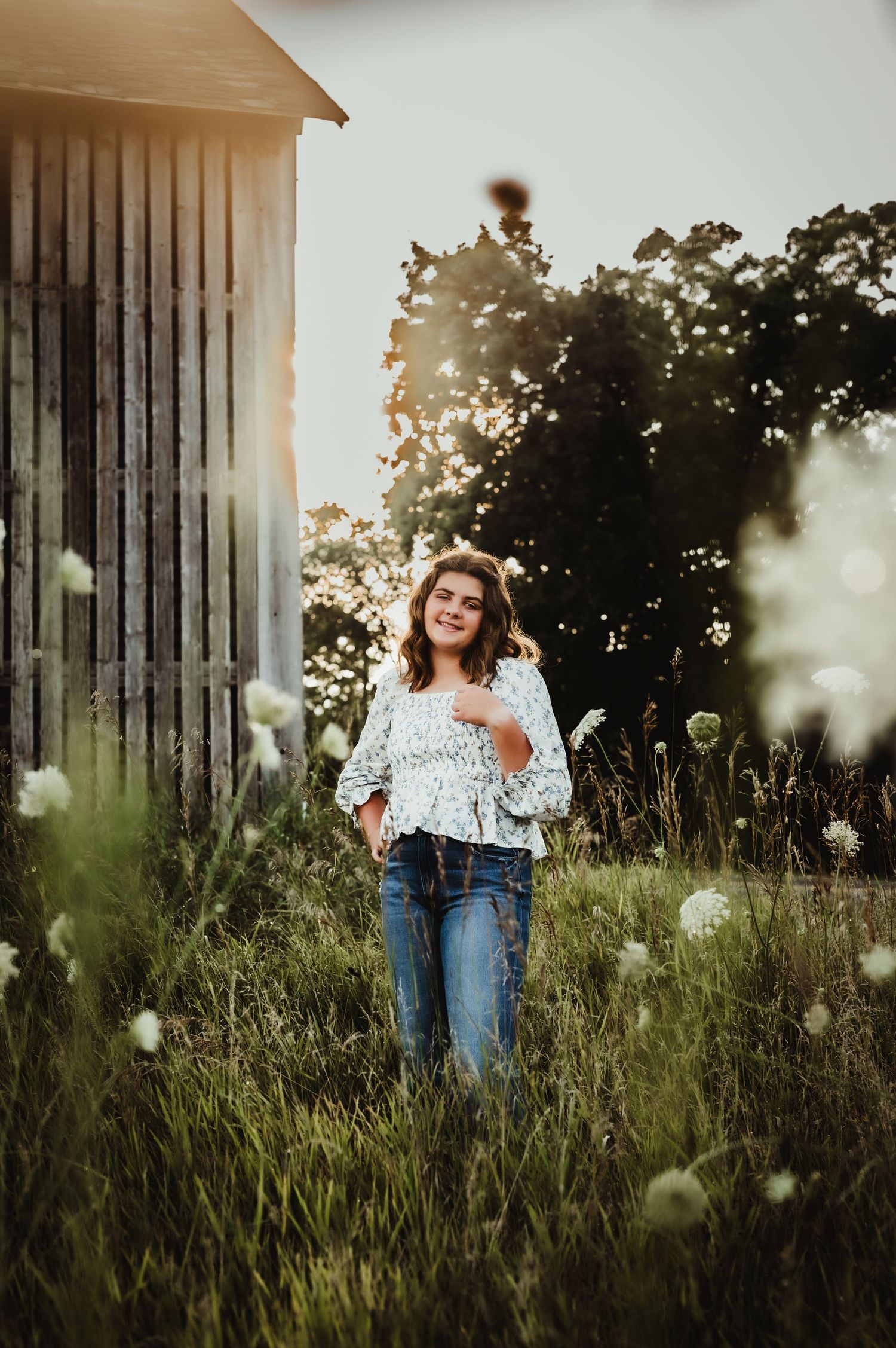 Portrait in tall grass near rustic barn with ethereal sunlight and dandelions.