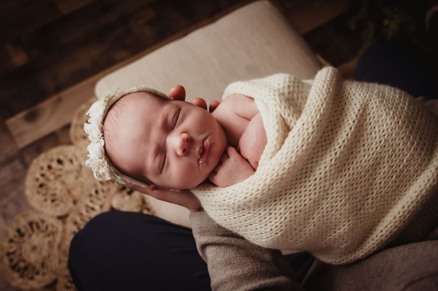Peaceful sleeping newborn wearing lace bonnet wrapped in cream knit blanket.