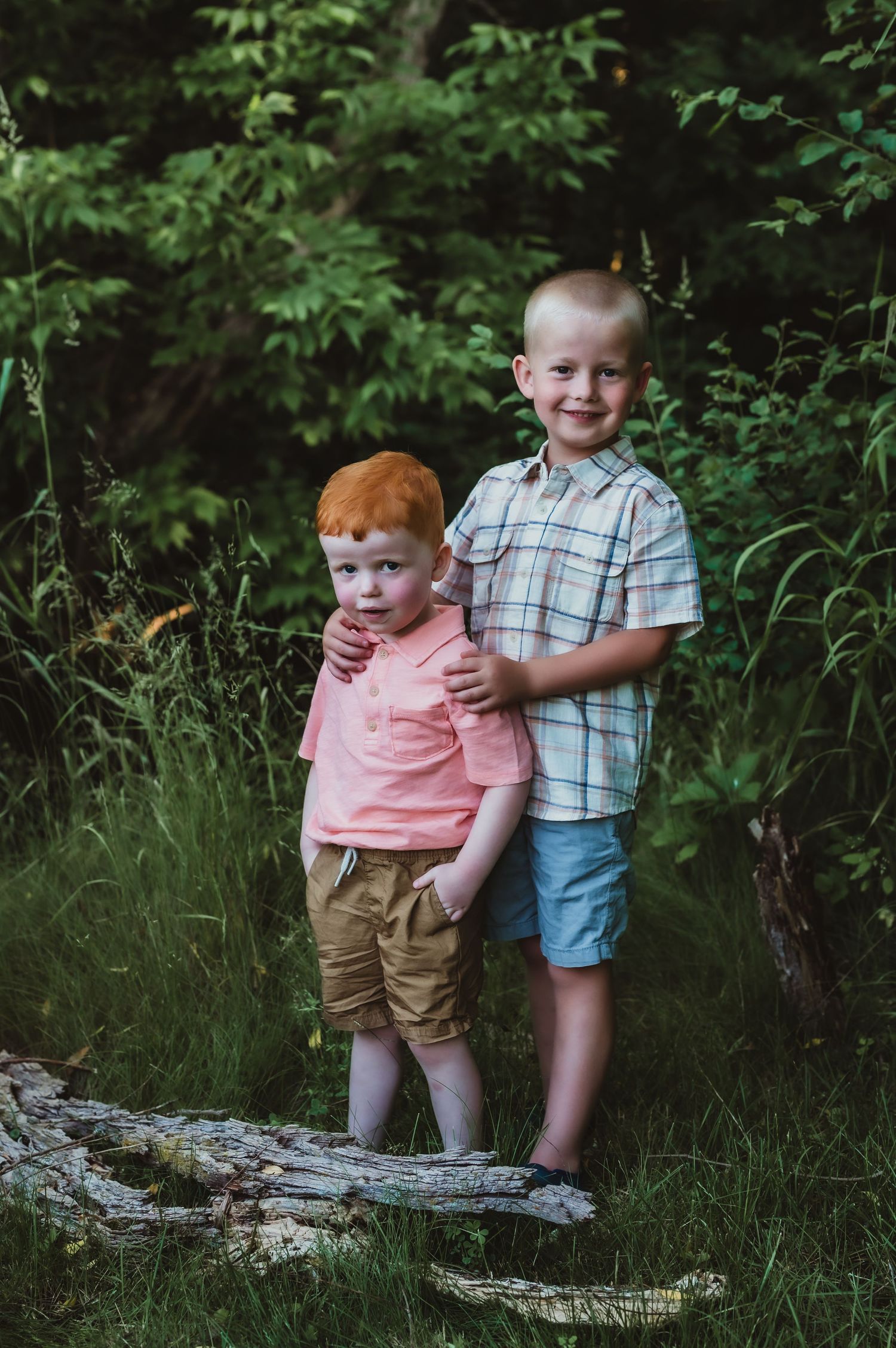 Two children standing together on a fallen log in a wooded area.