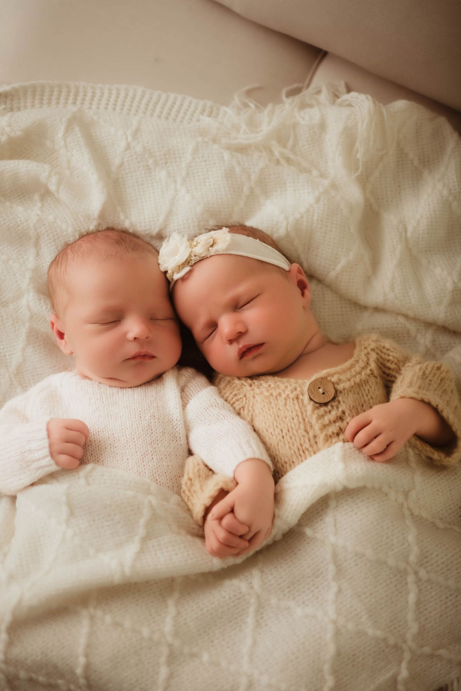 Two sleeping newborn babies in cozy knit outfits lie side by side on a soft cream blanket, one wearing a floral headband.