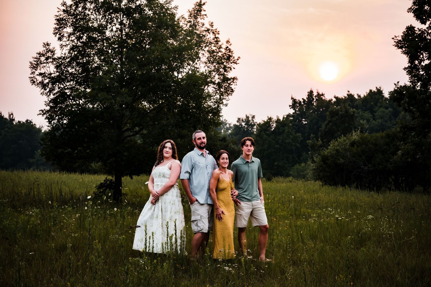Family of four posing in a sunlit meadow at sunset with lush green trees in the background.