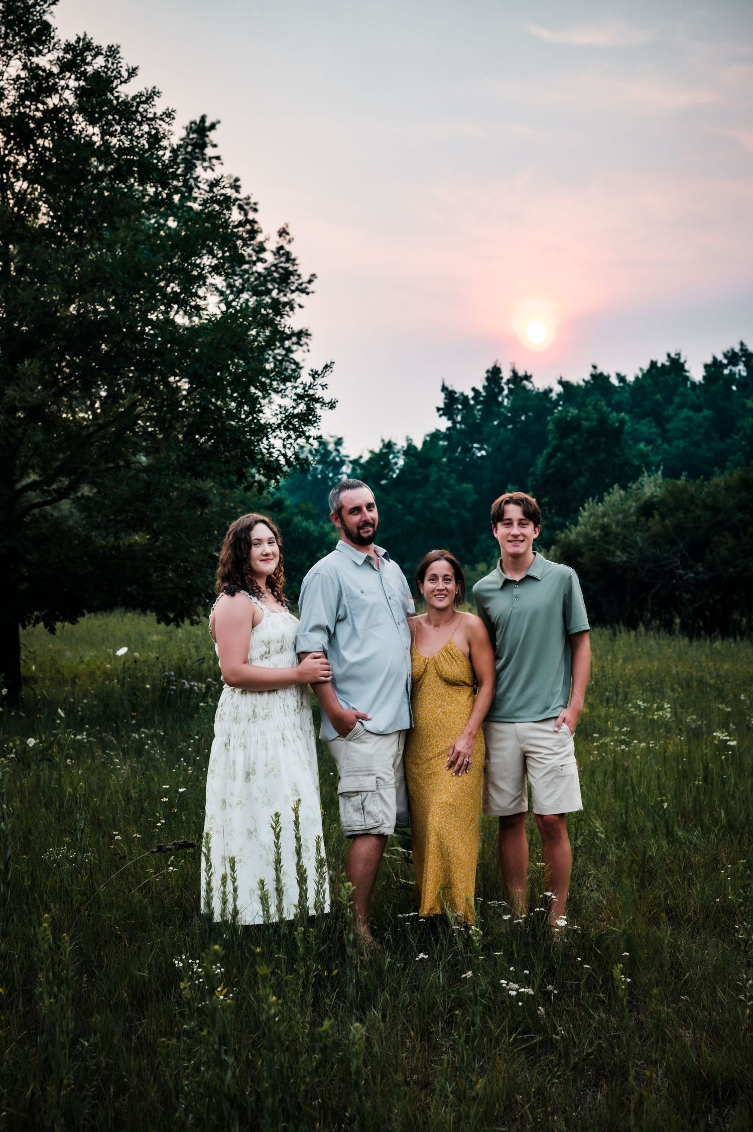 Family of four posing in a wildflower meadow at sunset, surrounded by lush green trees.