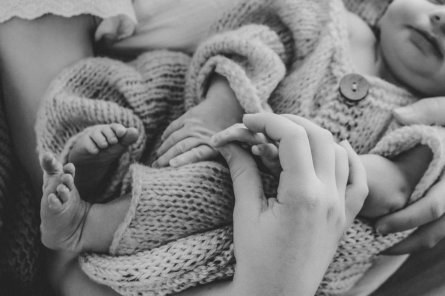 Black and white photo of a newborn baby held in a parent's arms, wearing a knit sweater, tiny feet and hands visible.