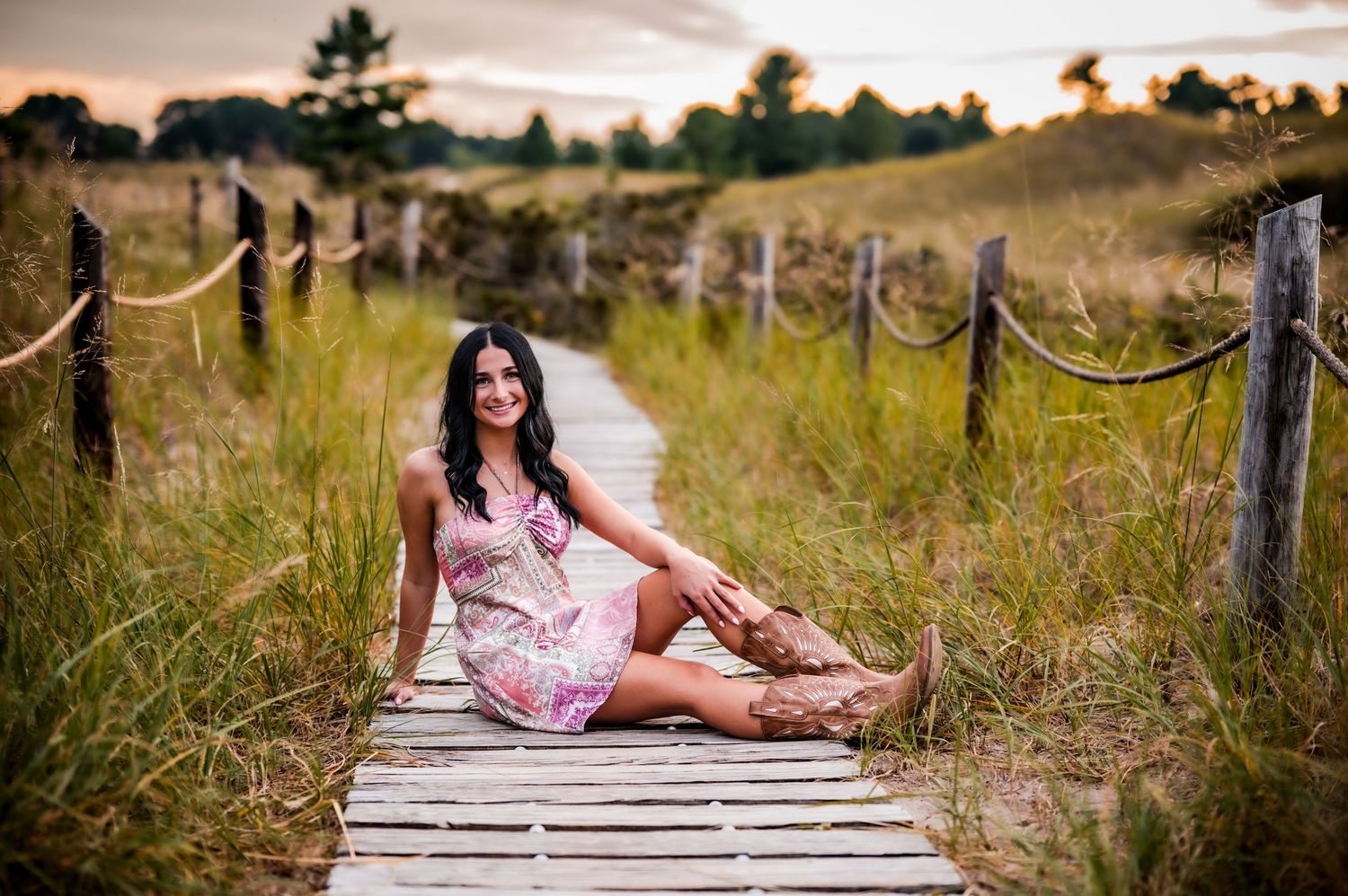A person in a floral dress poses on a wooden boardwalk surrounded by tall grass and natural scenery at sunset, Kewaskum, WI